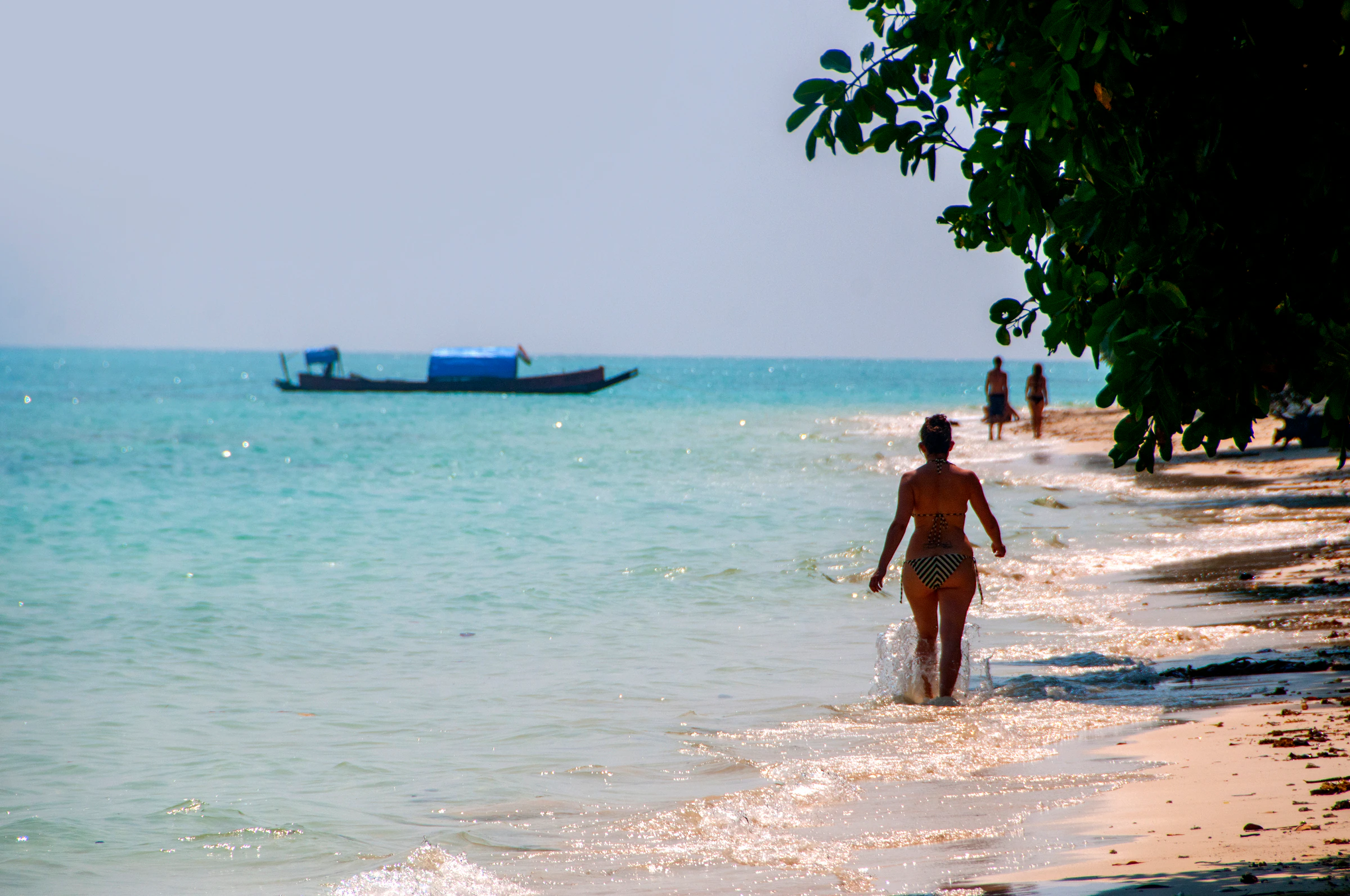 A woman walks through the clear shallows on a Havelock Island beach, with a fishing boat offshore