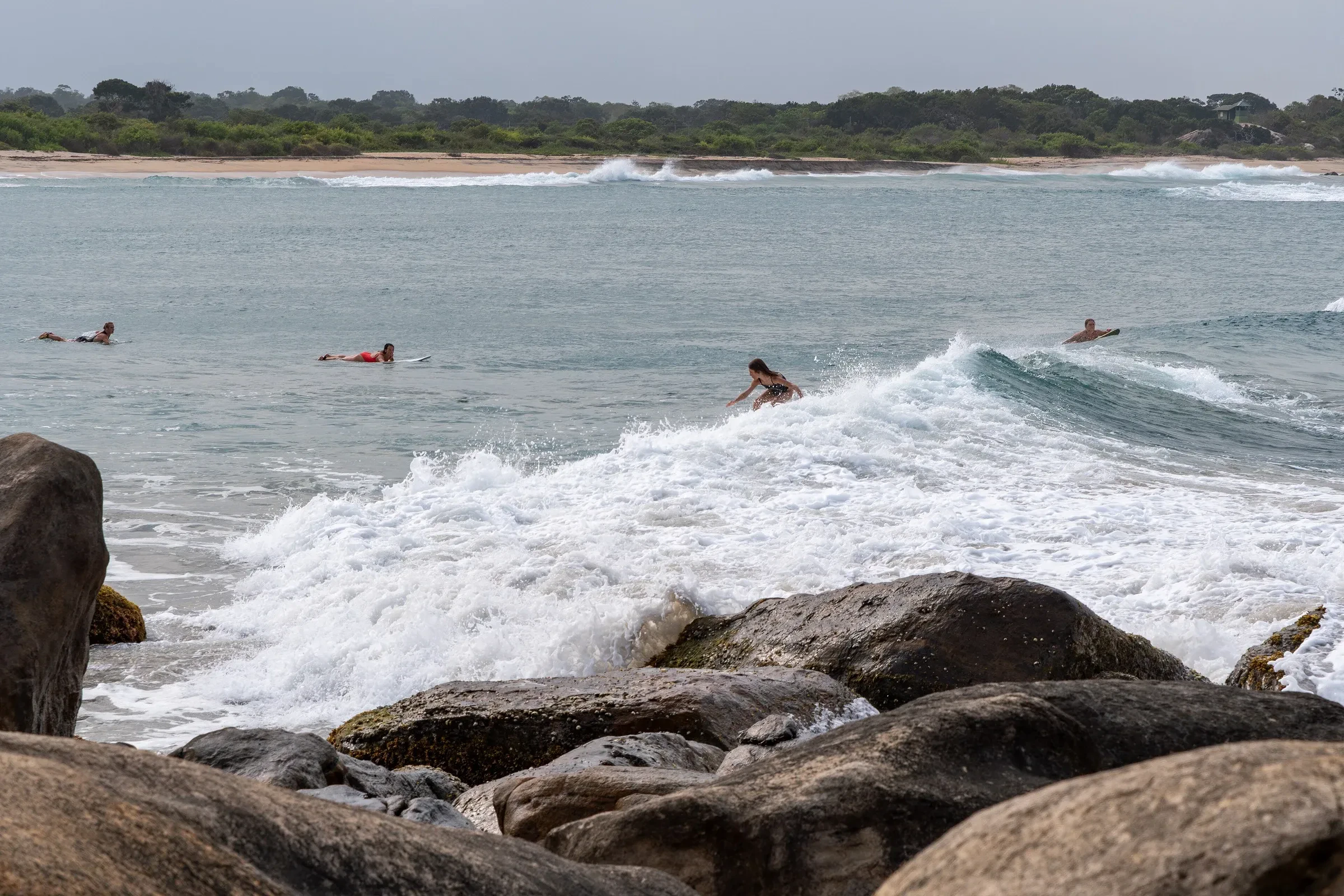 Surfers in the water beyond a rocky shore at Panama Beach, near Arugam Bay