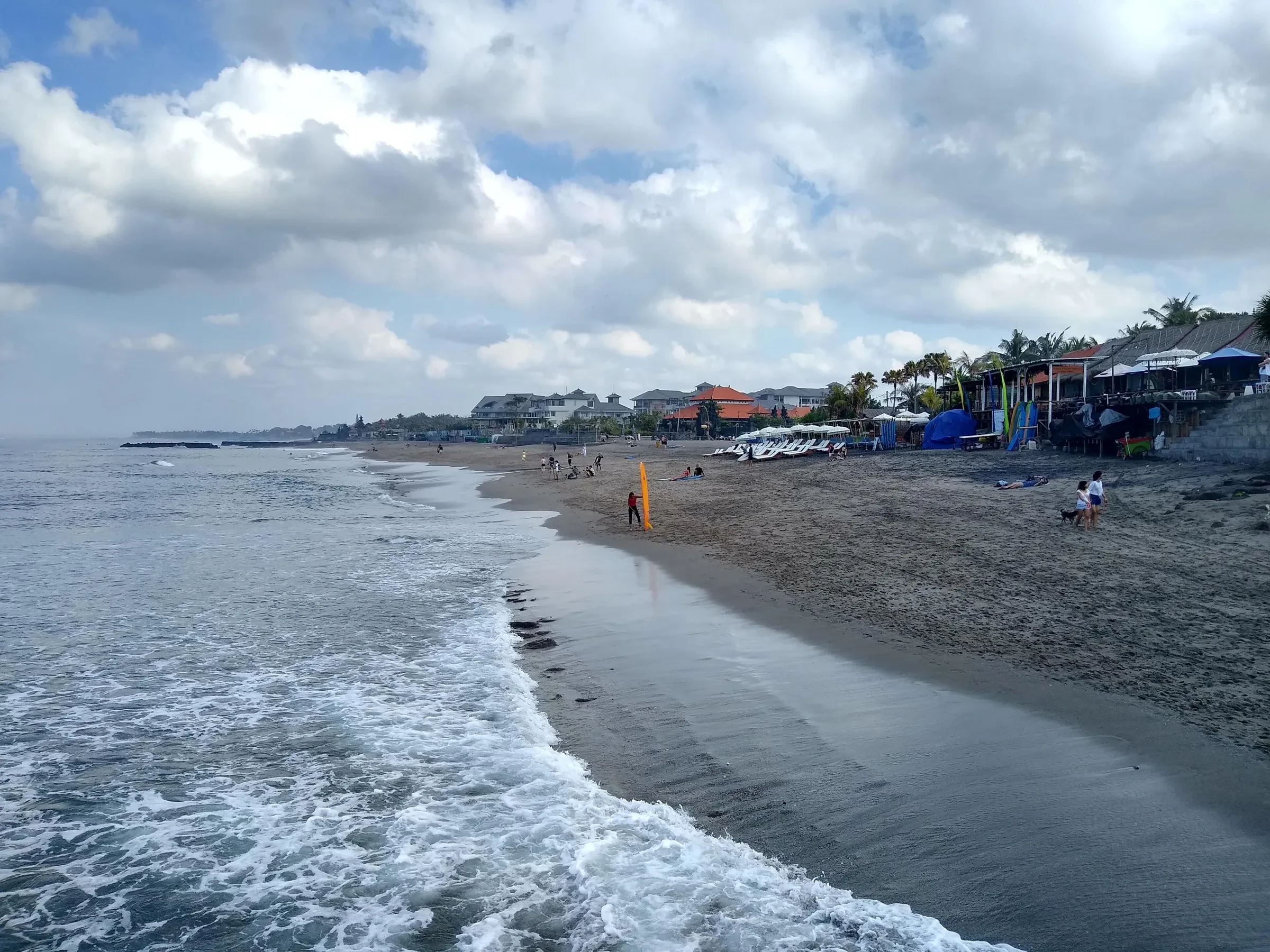 Waves rolling onto the dark sand of Canggu beach with beach cafes in the distance