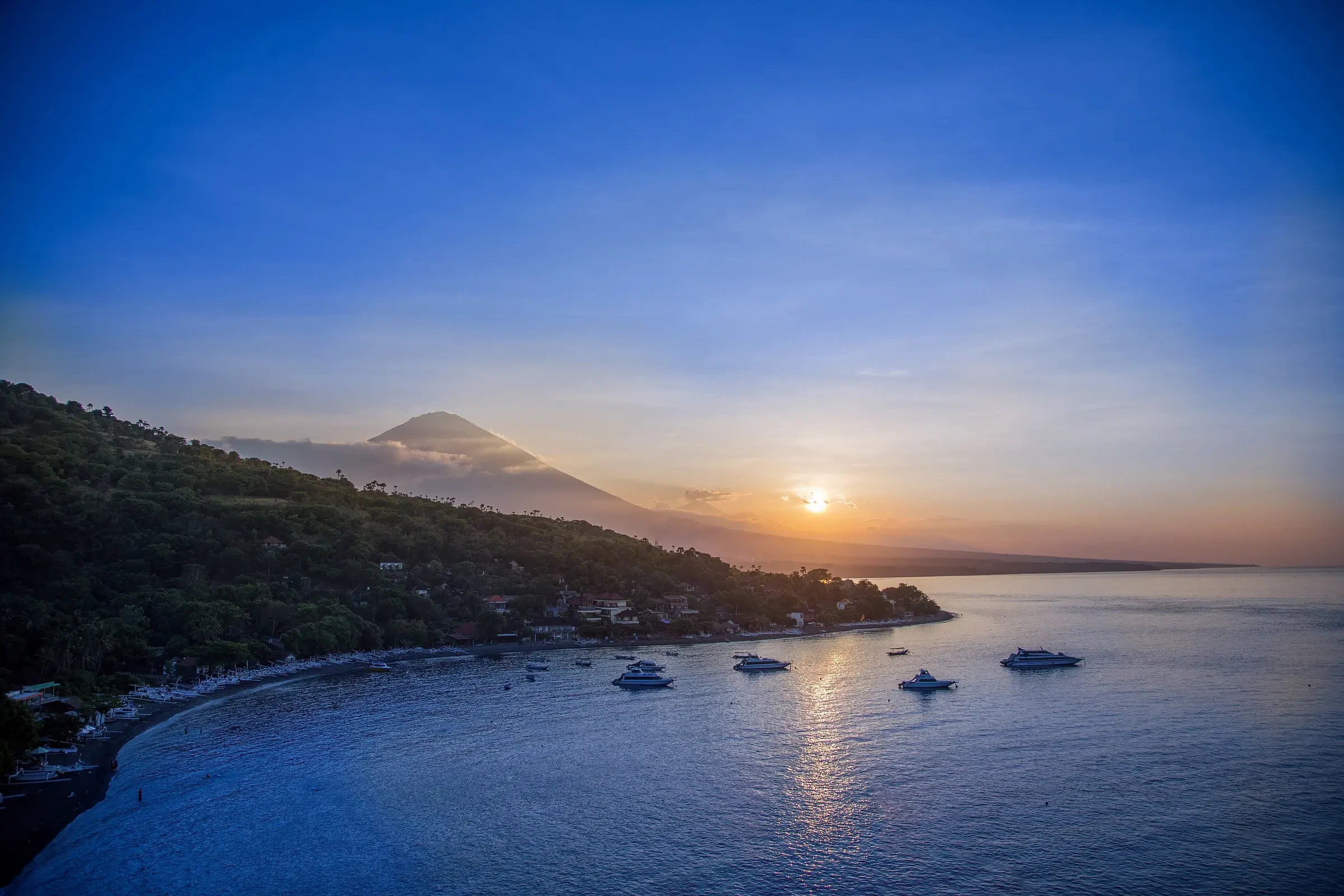 Amed's bay on Bali's east coast at sunset, with boats anchored offshore and Mount Agung on the horizon