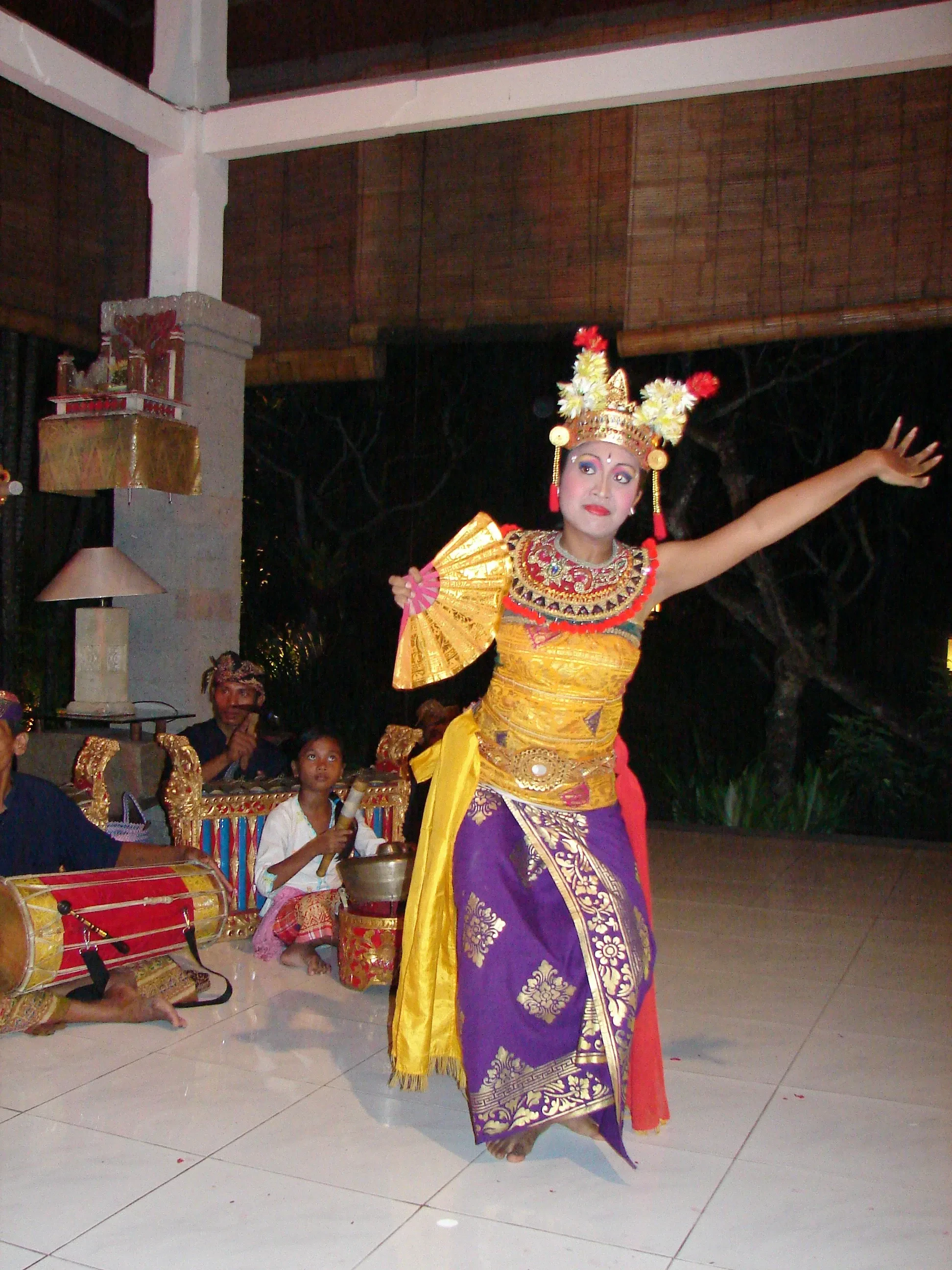 A young Legong dancer in a gold headdress performing with a gamelan behind her