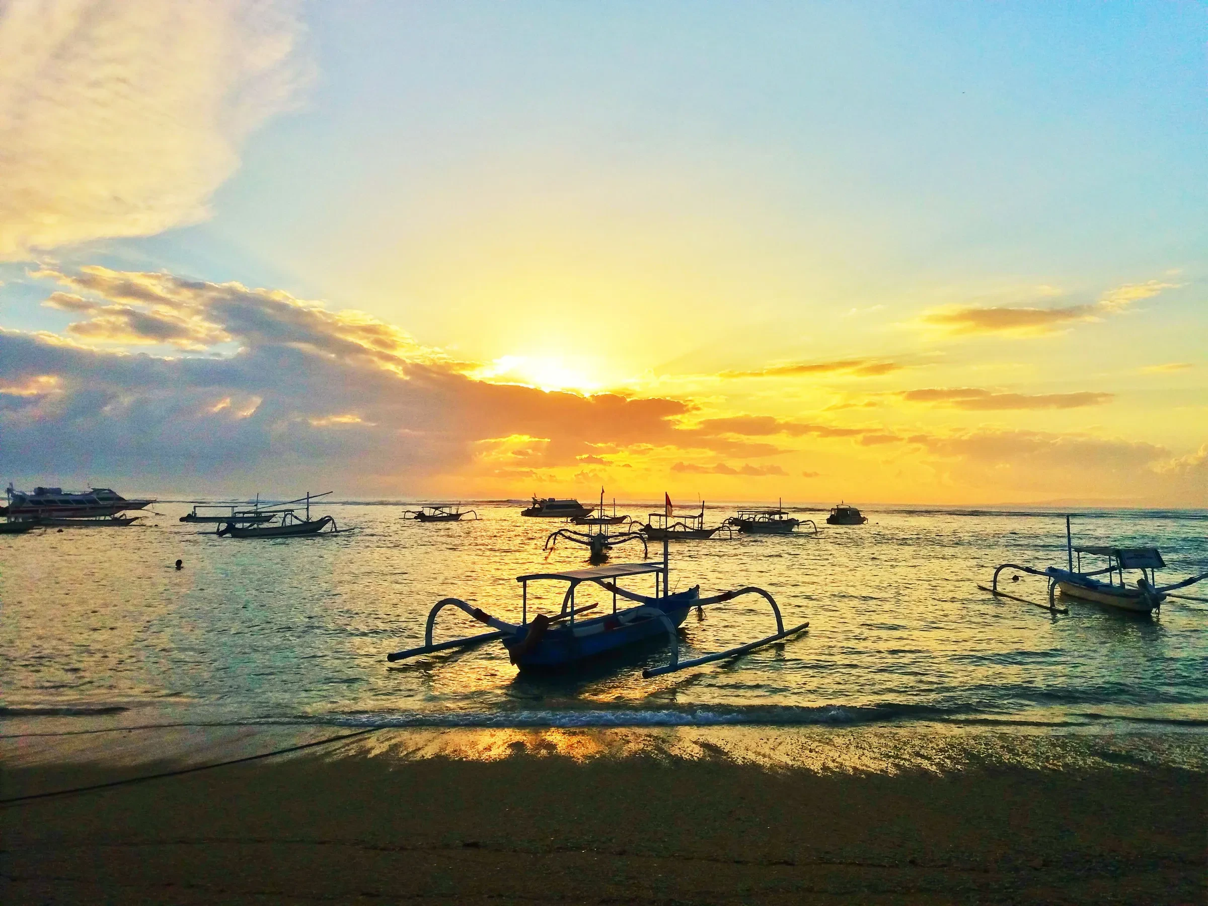 Outrigger fishing boats on calm water at Sanur beach, with a golden sky at sunrise