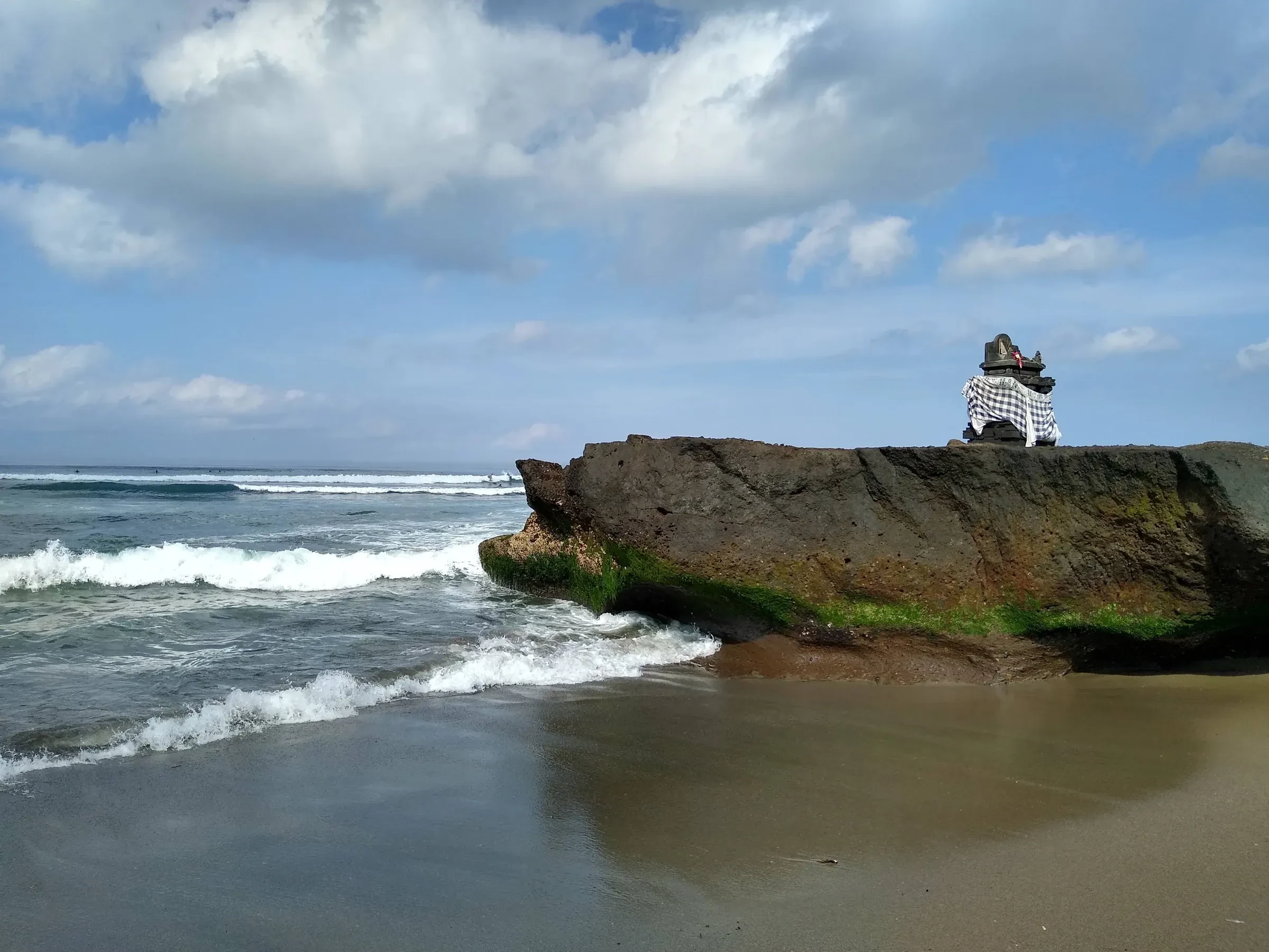 Waves rolling onto a large rock with a small altar on top, on the beach at Canggu