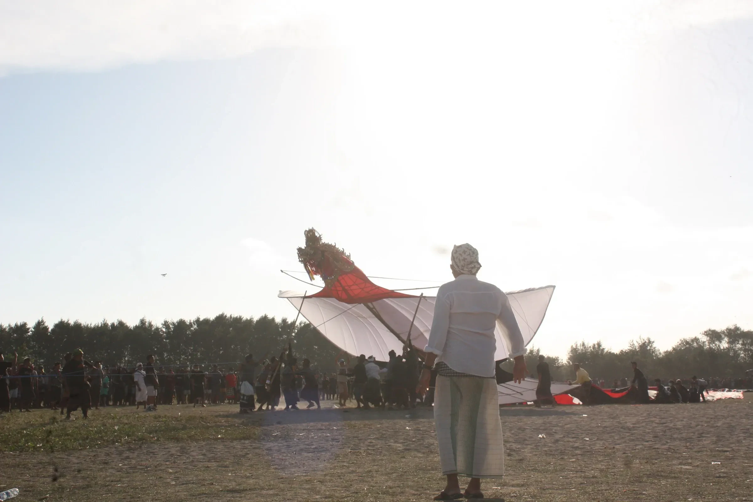 A man in white holds a large kite as a crowd gathers on Mertasari Beach during the kite festival in Sanur