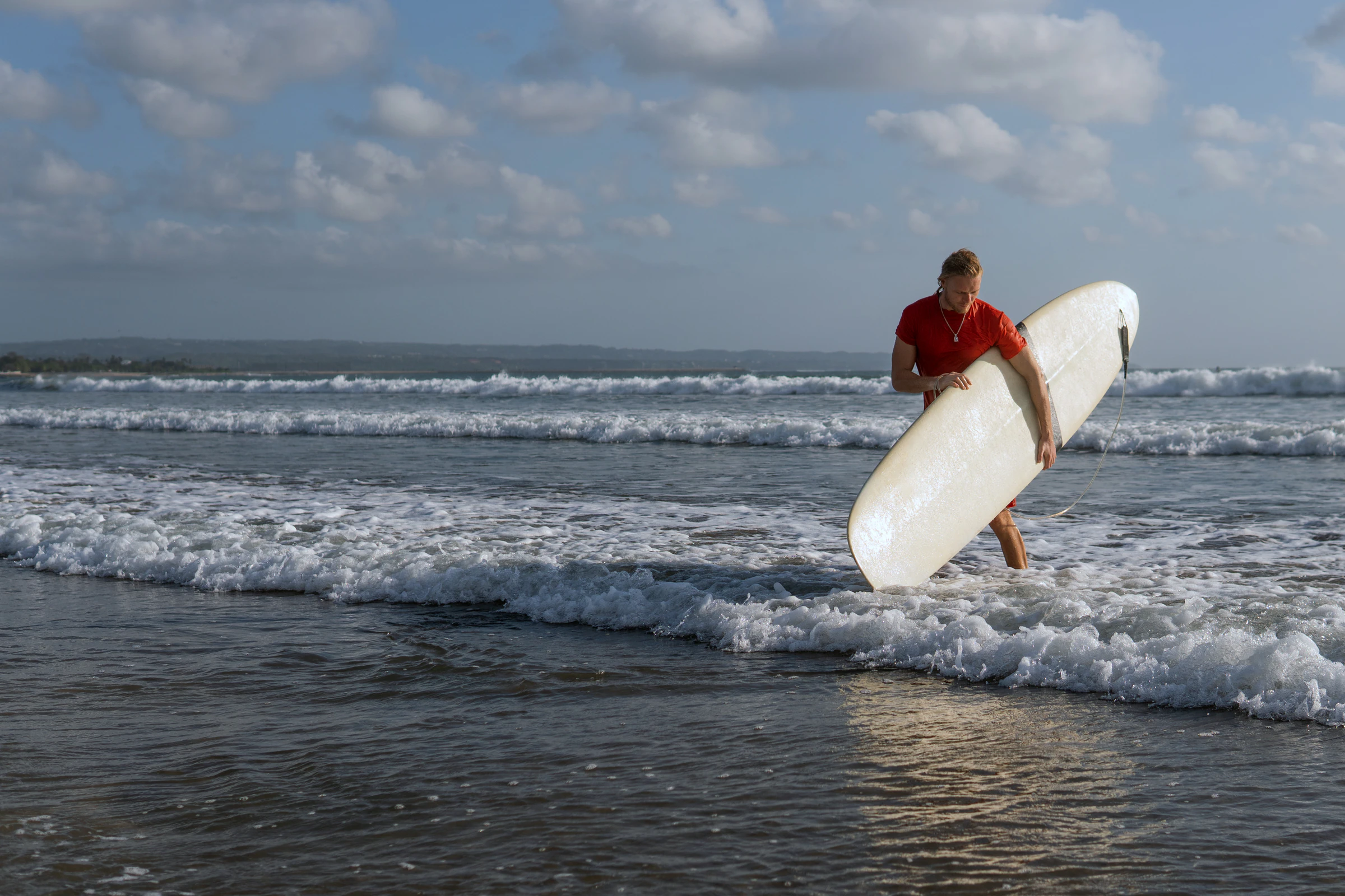 A surfer carrying a longboard out of the shallows on a Bali beach in daylight