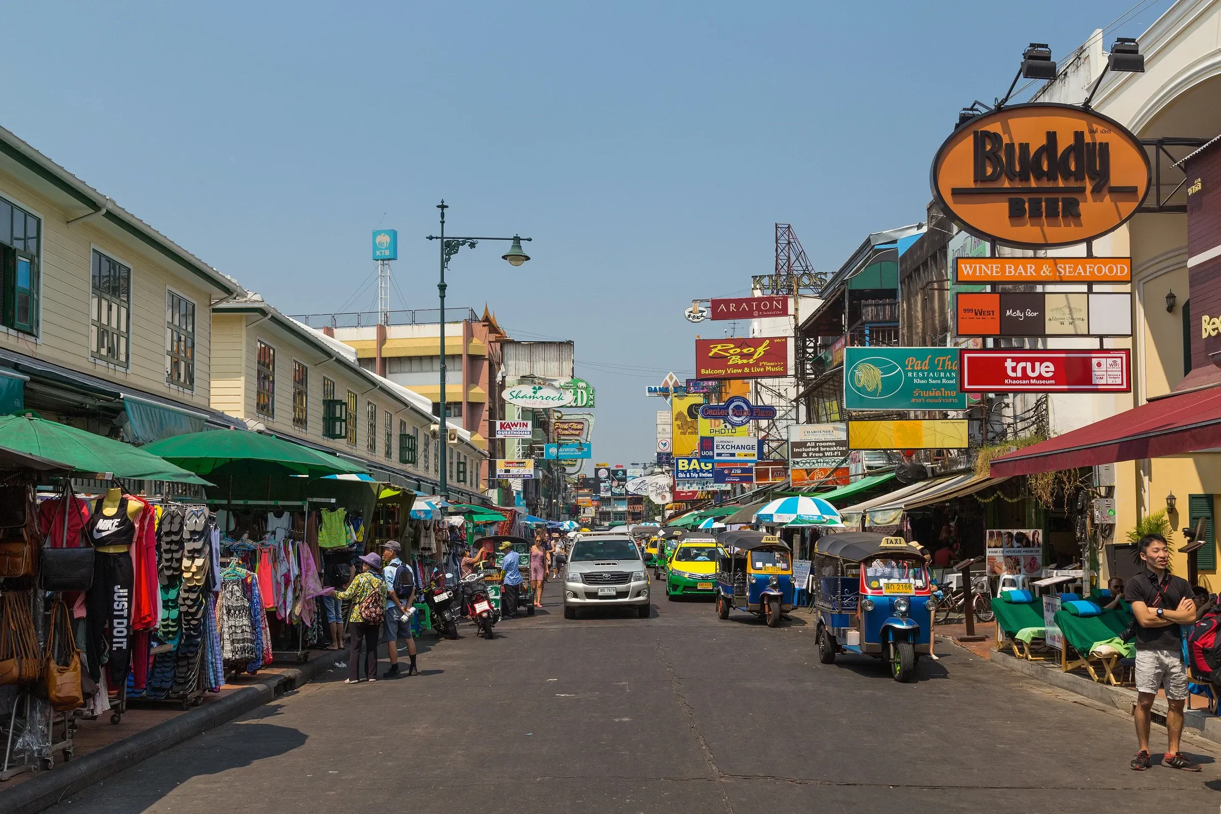 Khao San Road in daylight, with market stalls, shopfronts and a tuk-tuk on the street