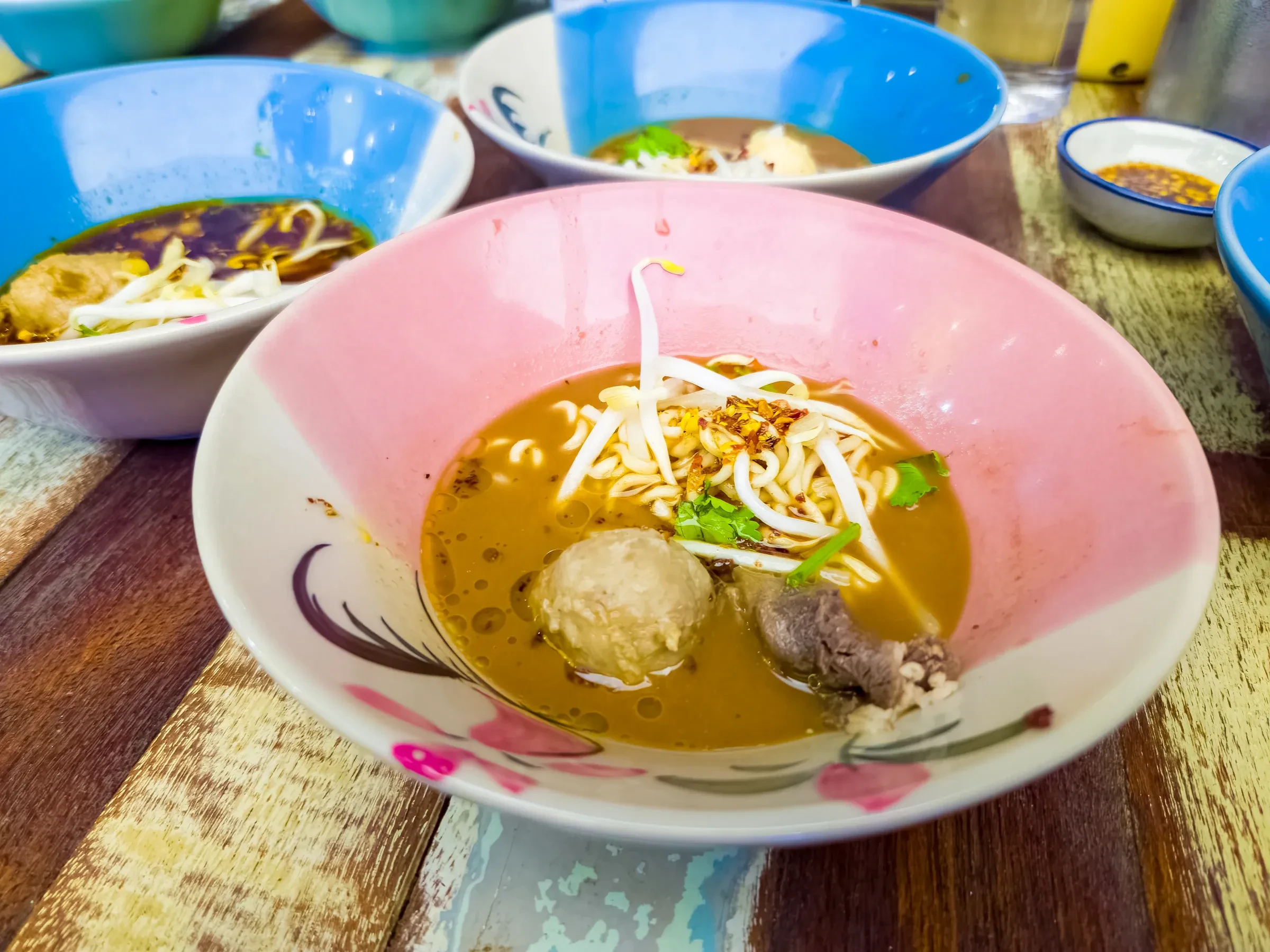 Bowls of Thai boat noodles with meatballs and bean sprouts on a wooden table