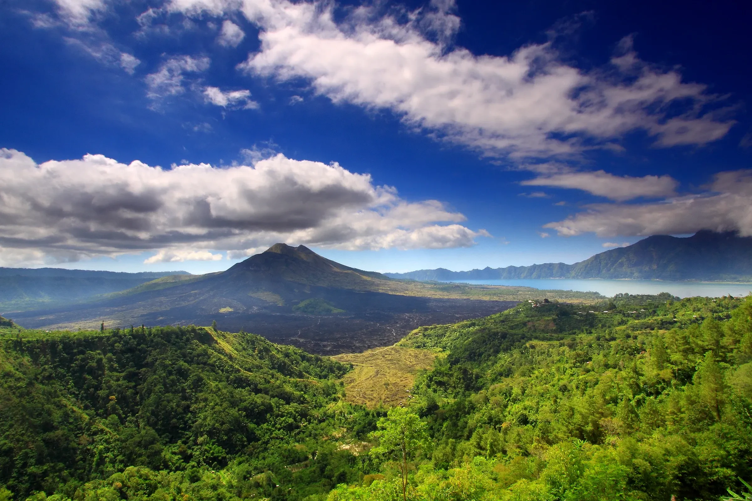 Mount Batur and its lake inside the caldera, seen from the south west under a blue sky with green hills in front.