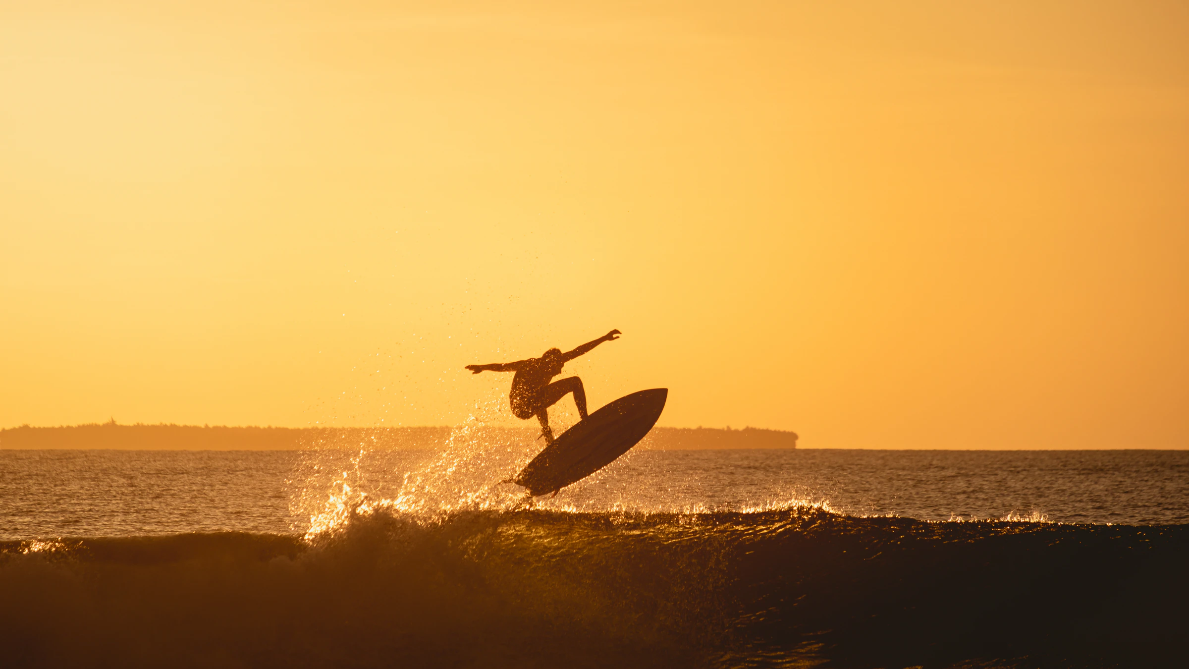 A surfer in silhouette launching off a wave against an orange sunset sky