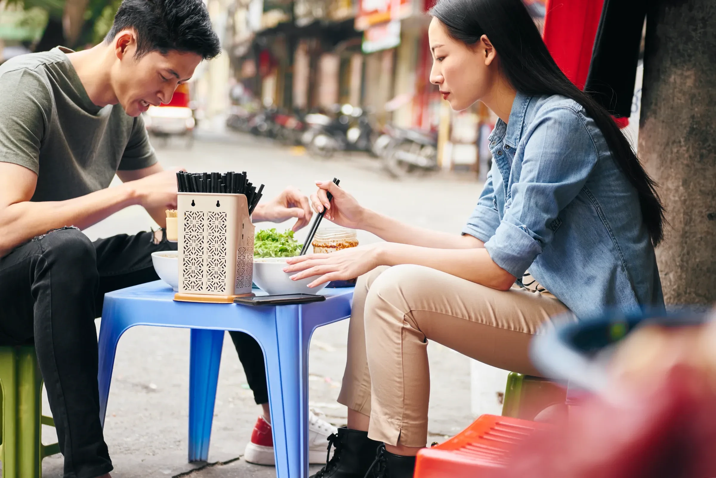 Two people eating street food at a low table on a Vietnamese street