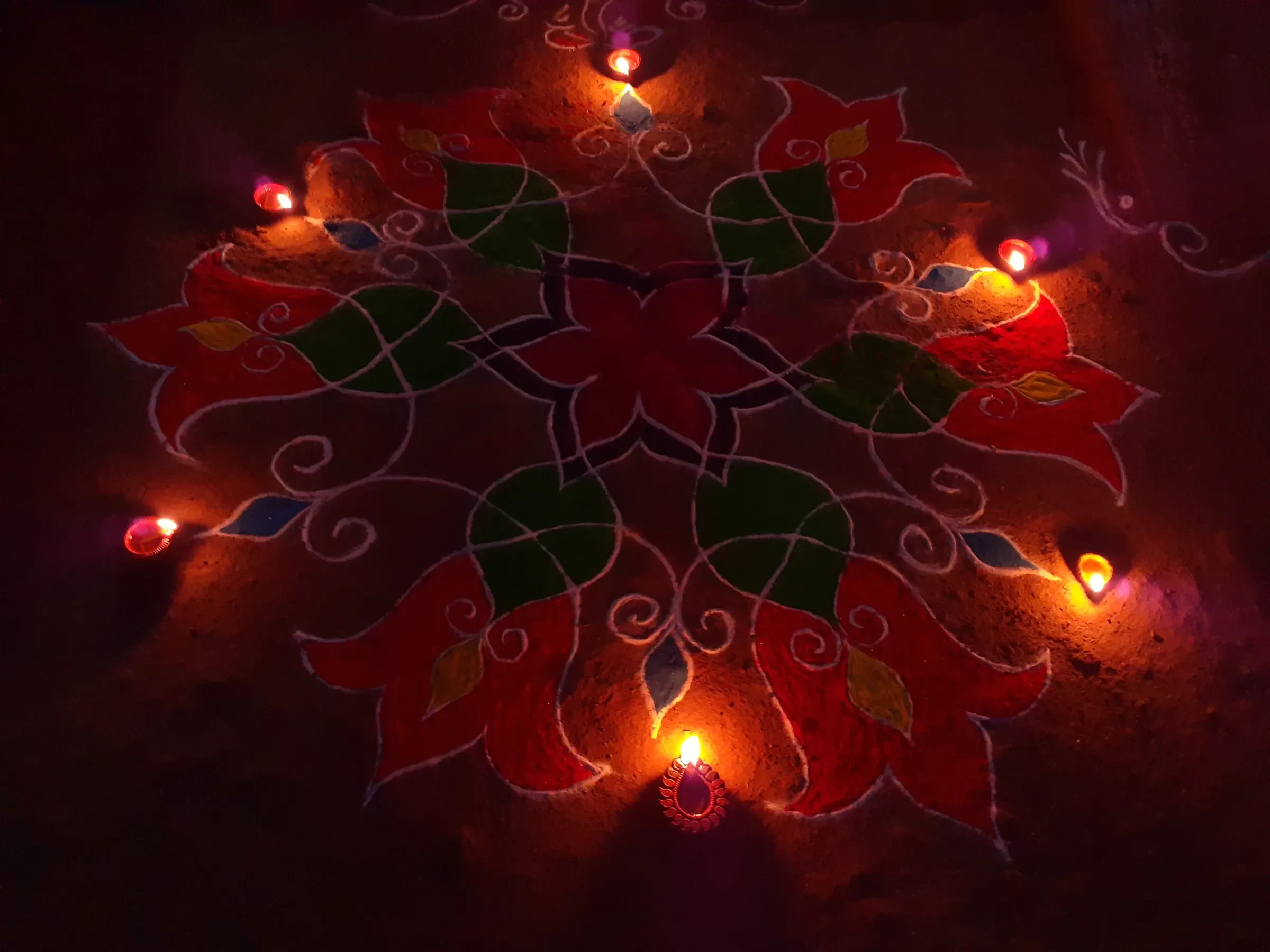 A Diwali rangoli in red and green with small clay lamps lit around it at night
