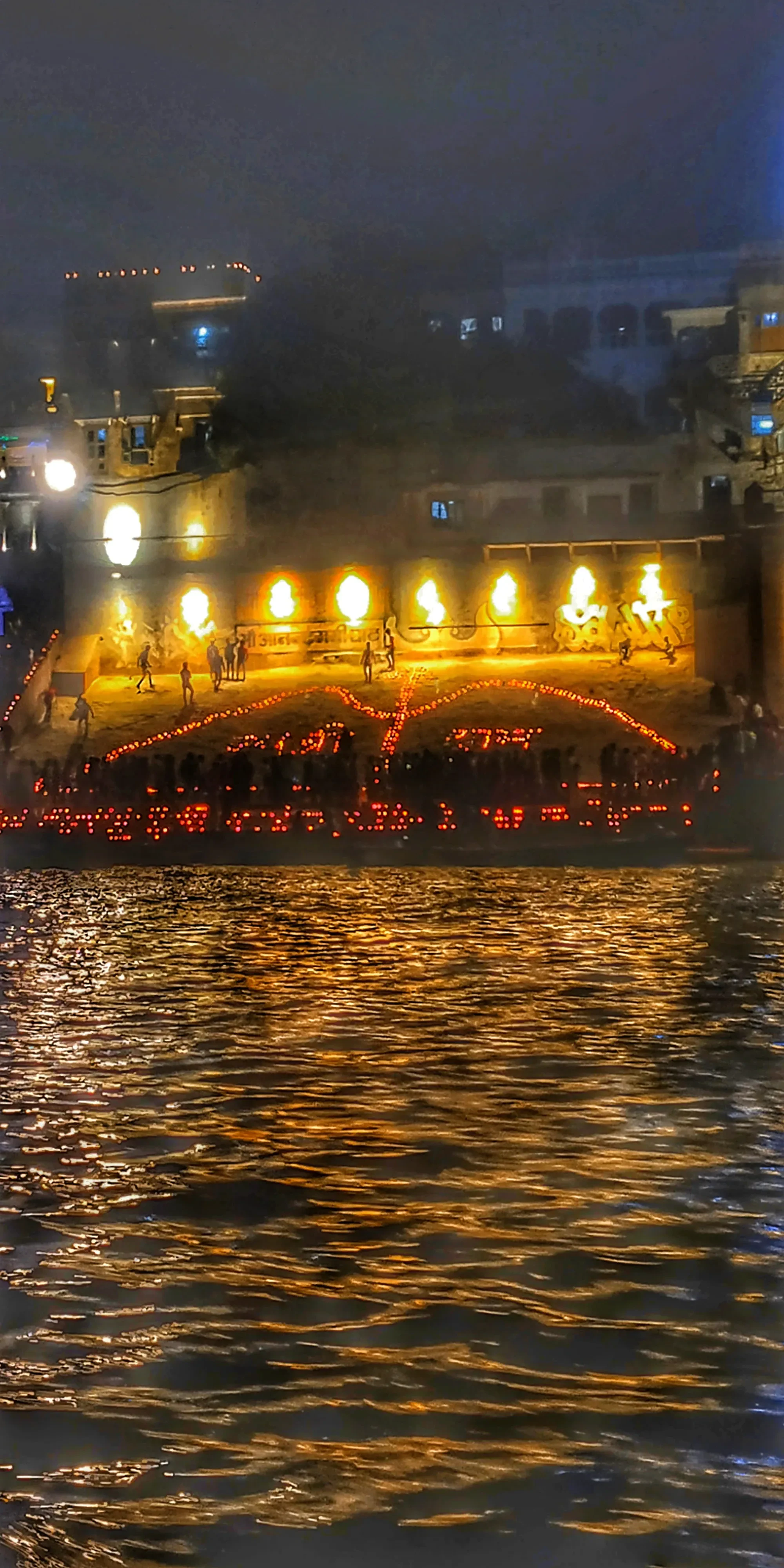 A lit ghat in Varanasi glowing gold and reflected in the Ganges at night during Dev Diwali