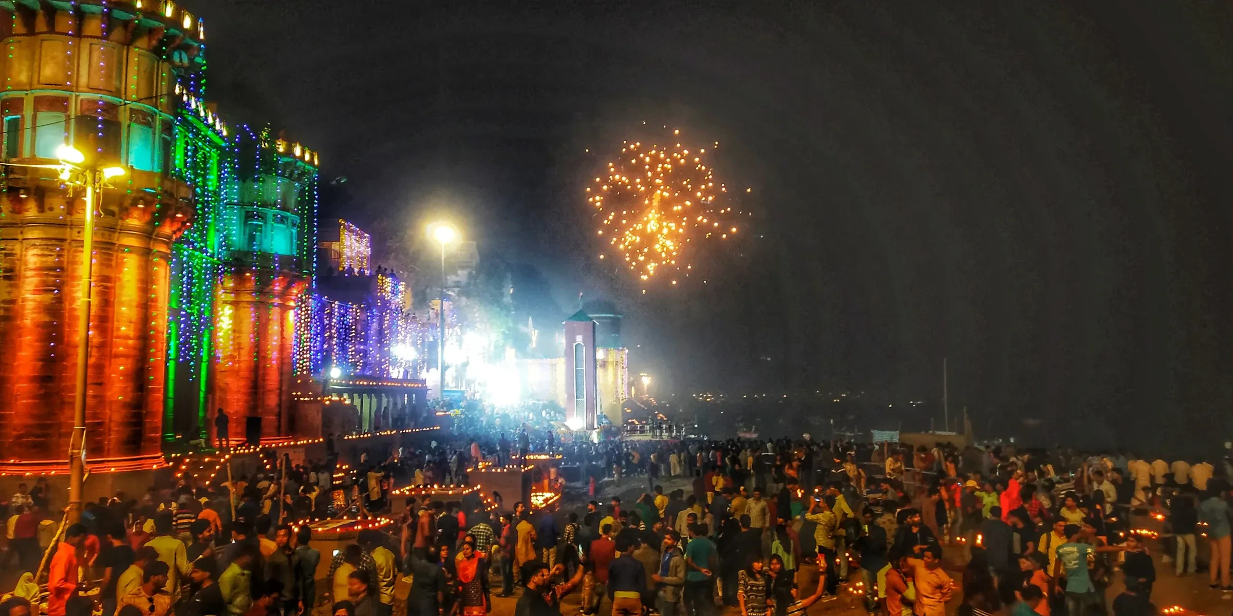 Colourfully lit ghats and a burst of fireworks above the crowd on the riverbank at Dev Diwali in Varanasi