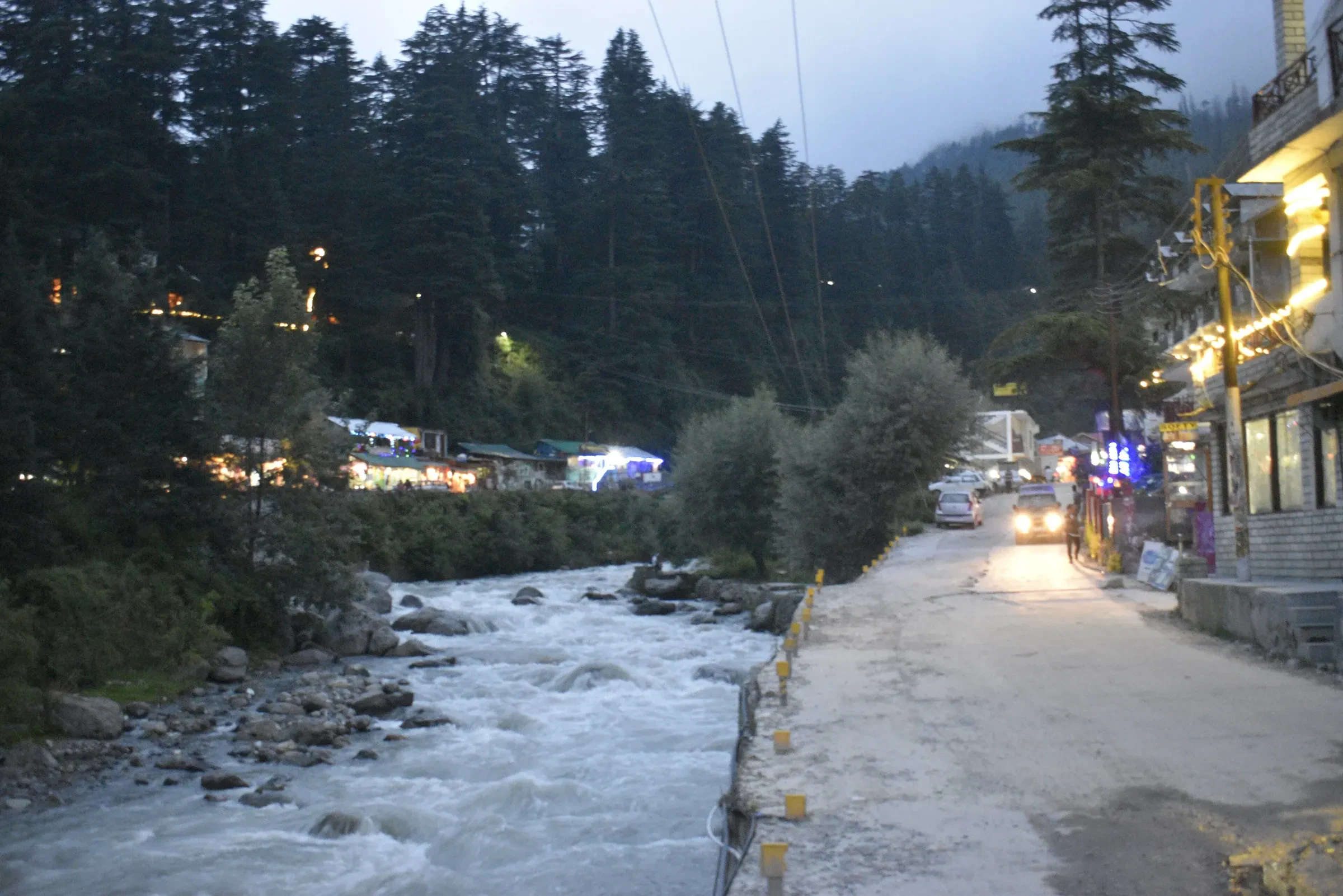 A riverside street in Old Manali at dusk, with pine forest behind the guesthouses