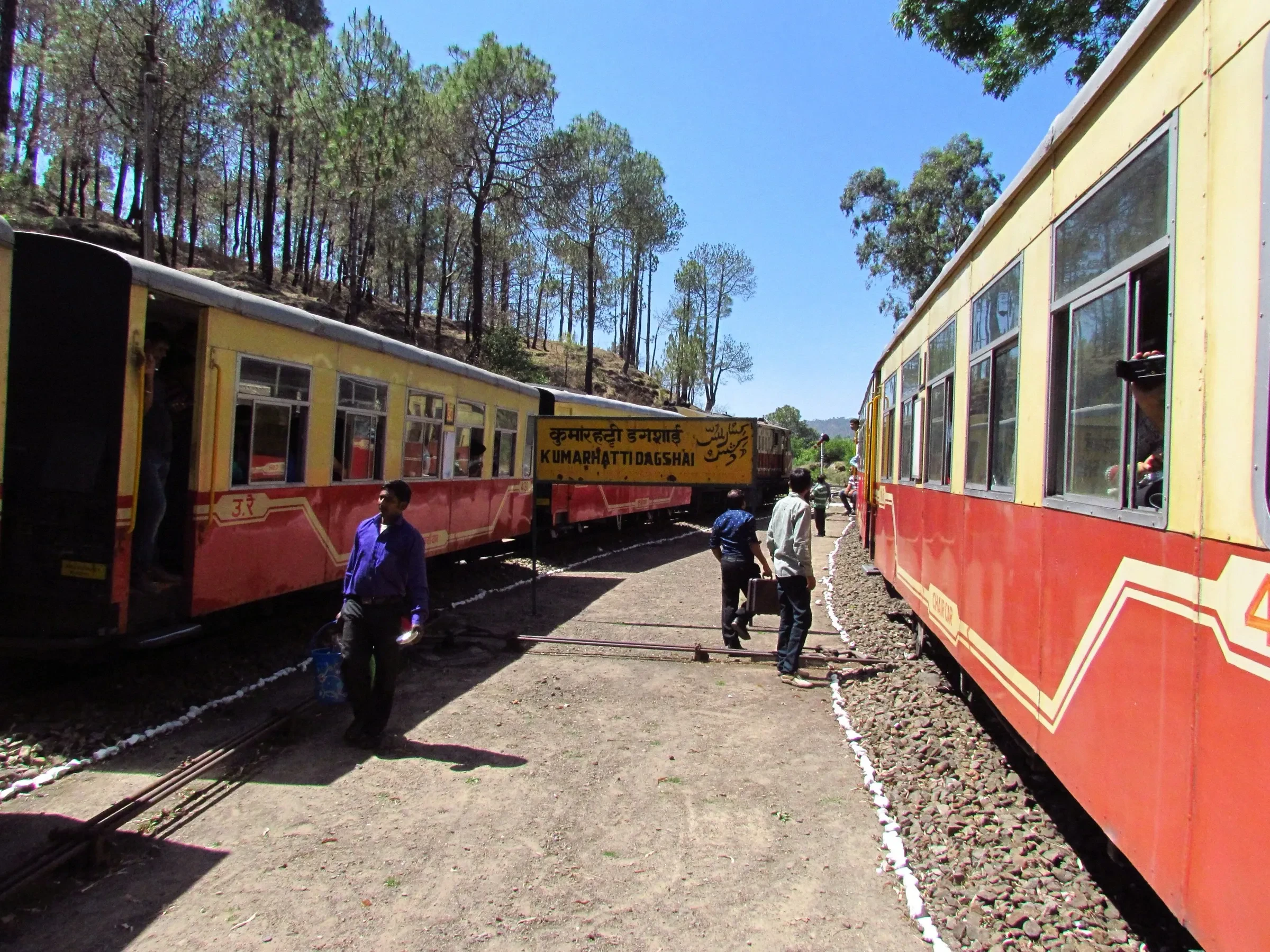A red and yellow Kalka-Shimla toy train waiting at a hill platform among pine trees