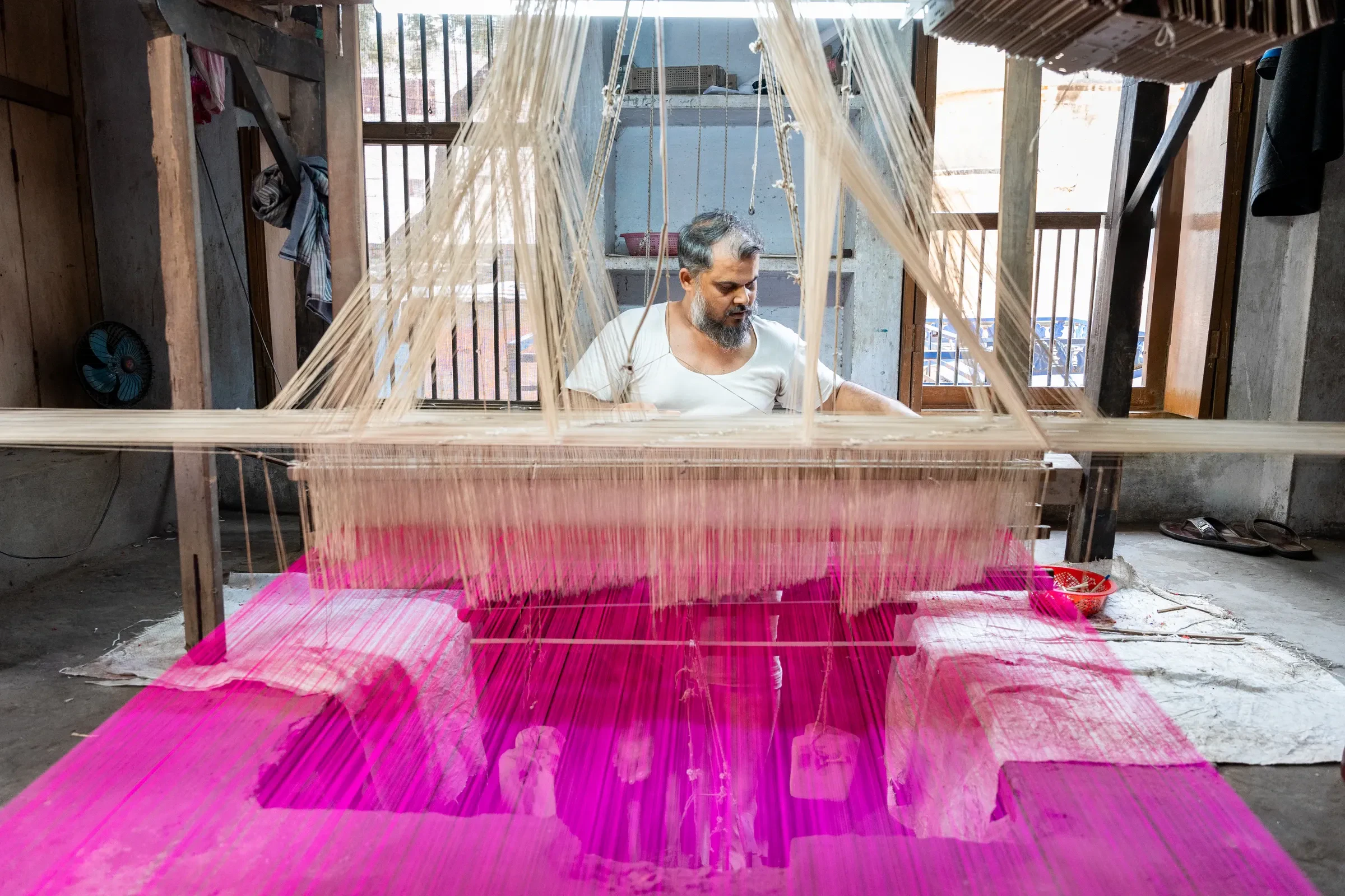 A weaver sitting at a handloom strung with bright pink thread