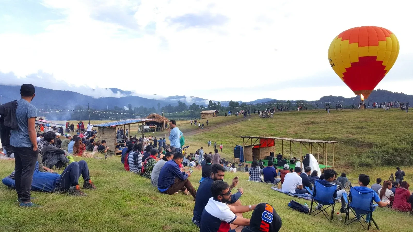 Festival-goers sitting on the grass at the Ziro festival ground with mountains behind and a hot-air balloon overhead