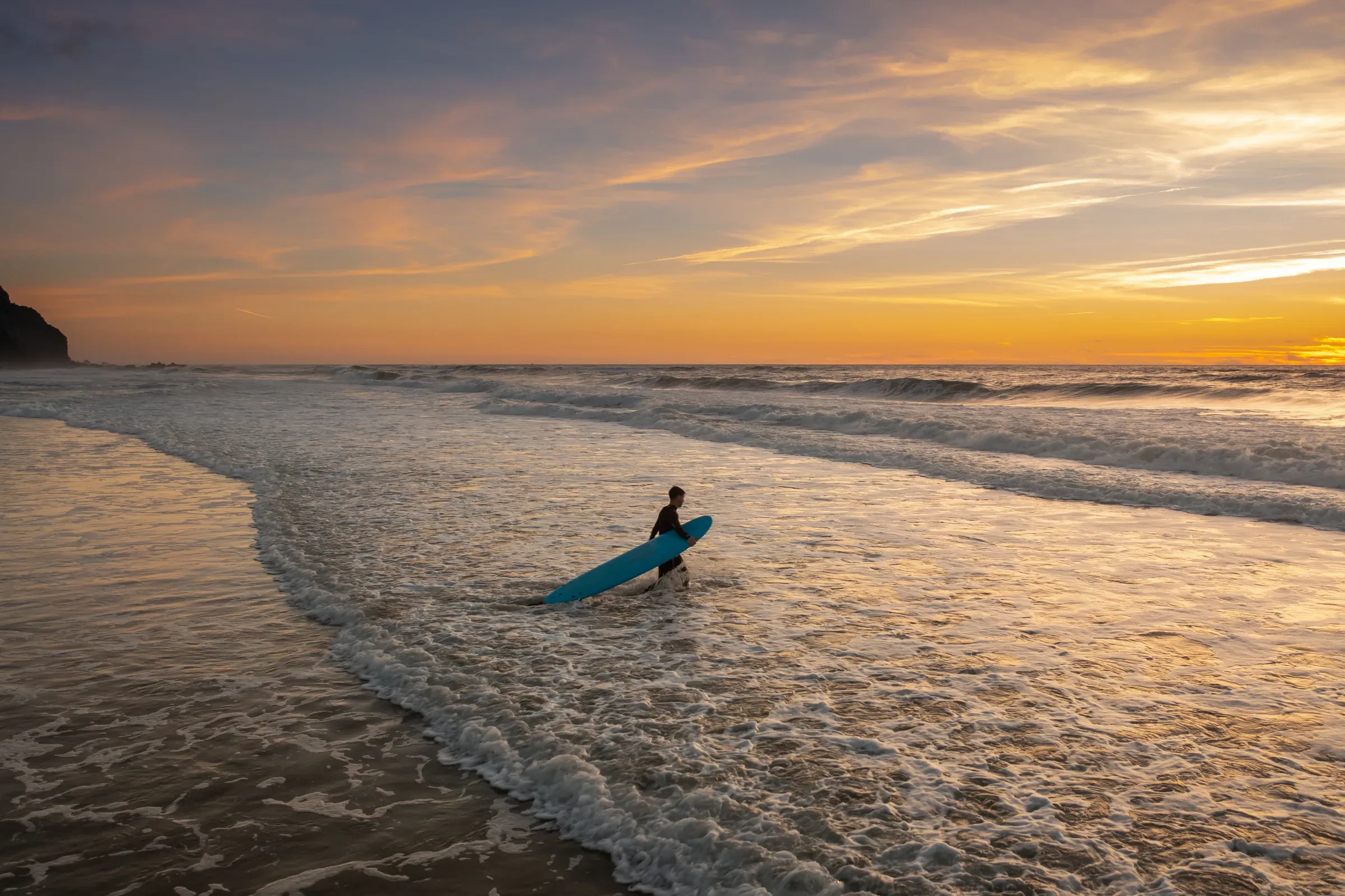 A surfer walking down to the sea with a board at sunset