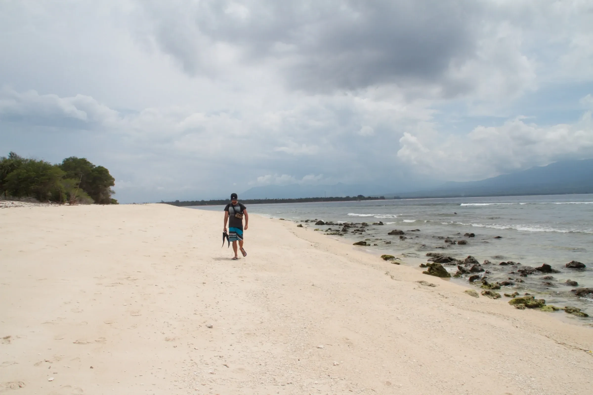 A walker on a pale sand beach on Gili Air under cloudy skies