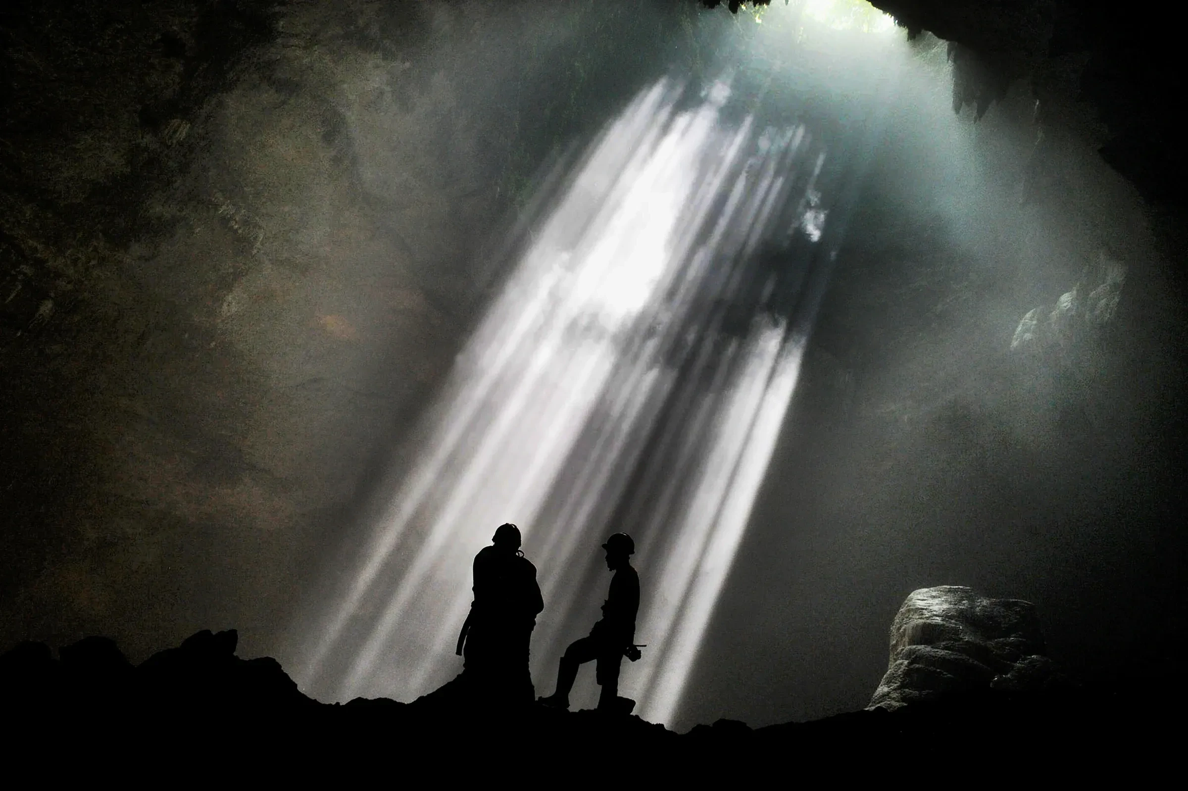 Beams of light falling into Jomblang Cave, Gunung Kidul, with two silhouetted visitors