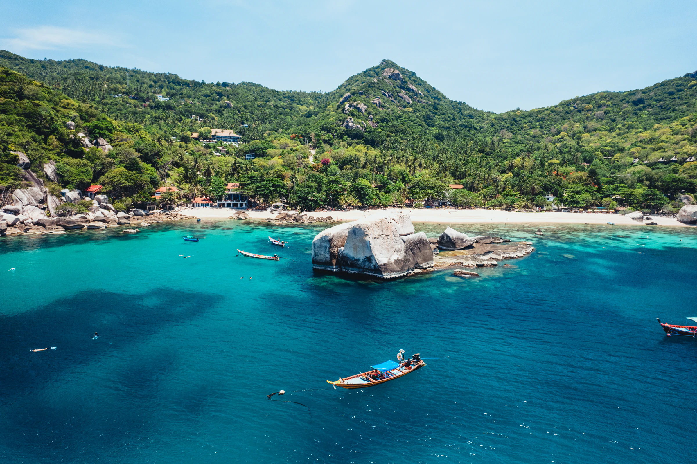 A longtail boat on clear turquoise water in a bay on Koh Tao, with the island's green hills behind