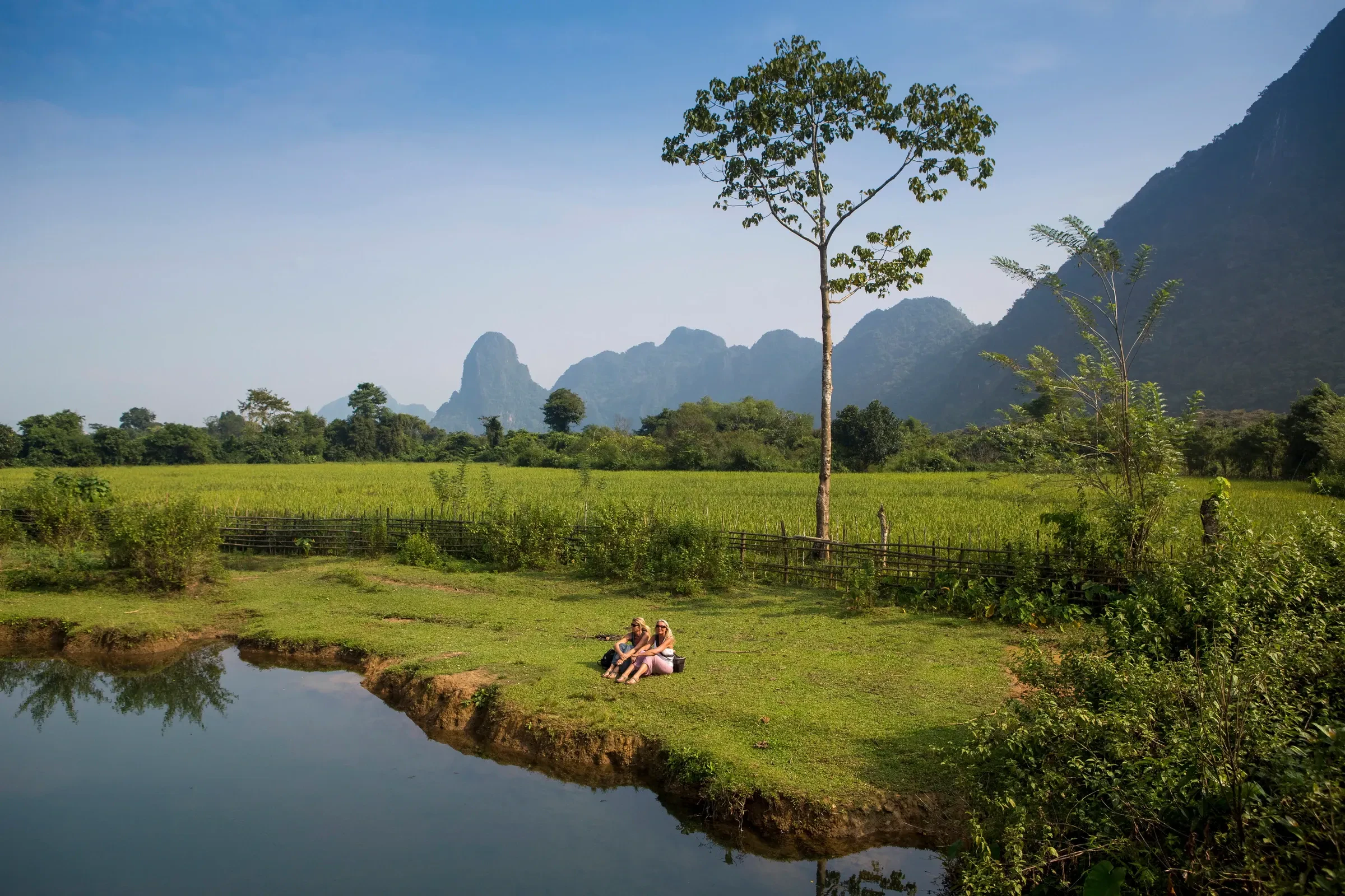 Two people sitting on a grassy bank beside the Nam Song, with limestone peaks behind, near Vang Vieng