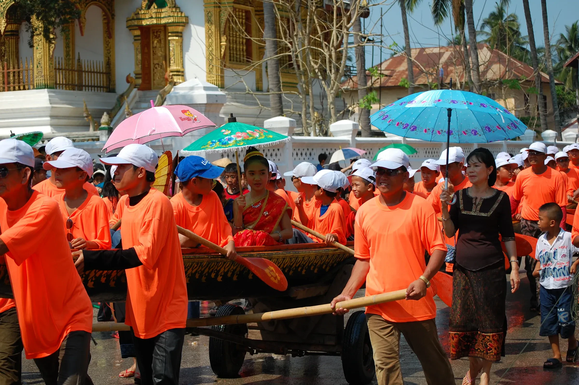A Lao New Year parade with people in orange shirts pulling a decorated river boat float along a street