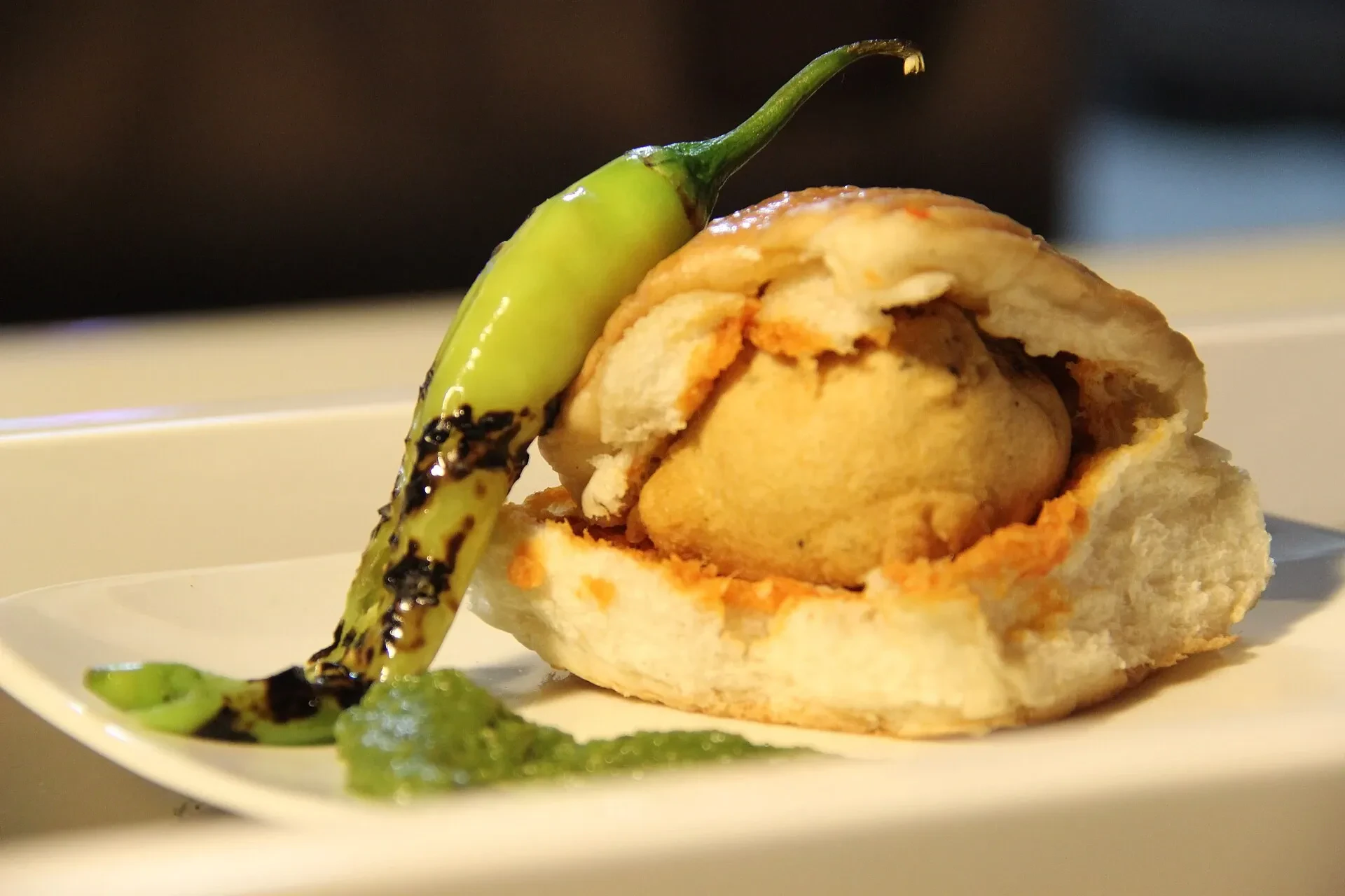 A vada pav, a spiced potato fritter in a soft bread roll, served on a plate with a green chilli