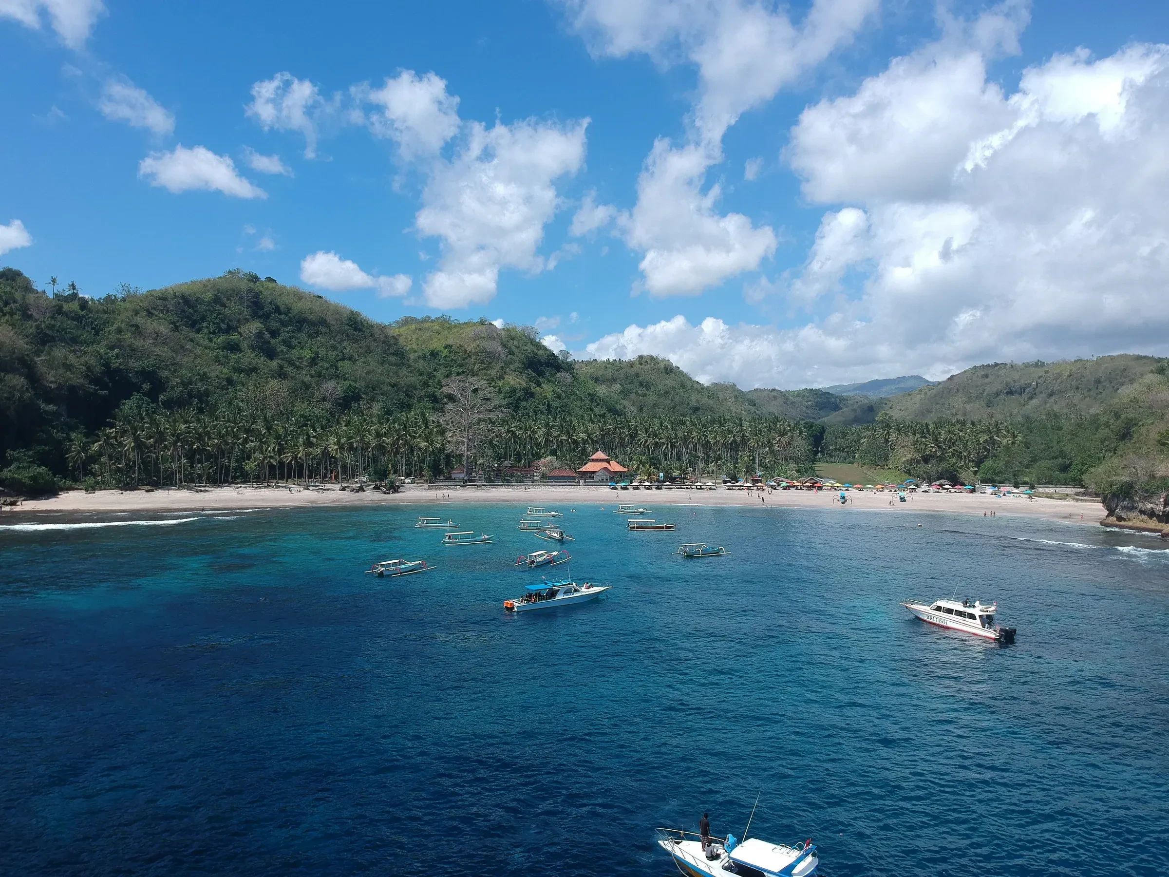 Boats anchored in the blue water of Crystal Bay, with a palm-lined beach and green hills behind