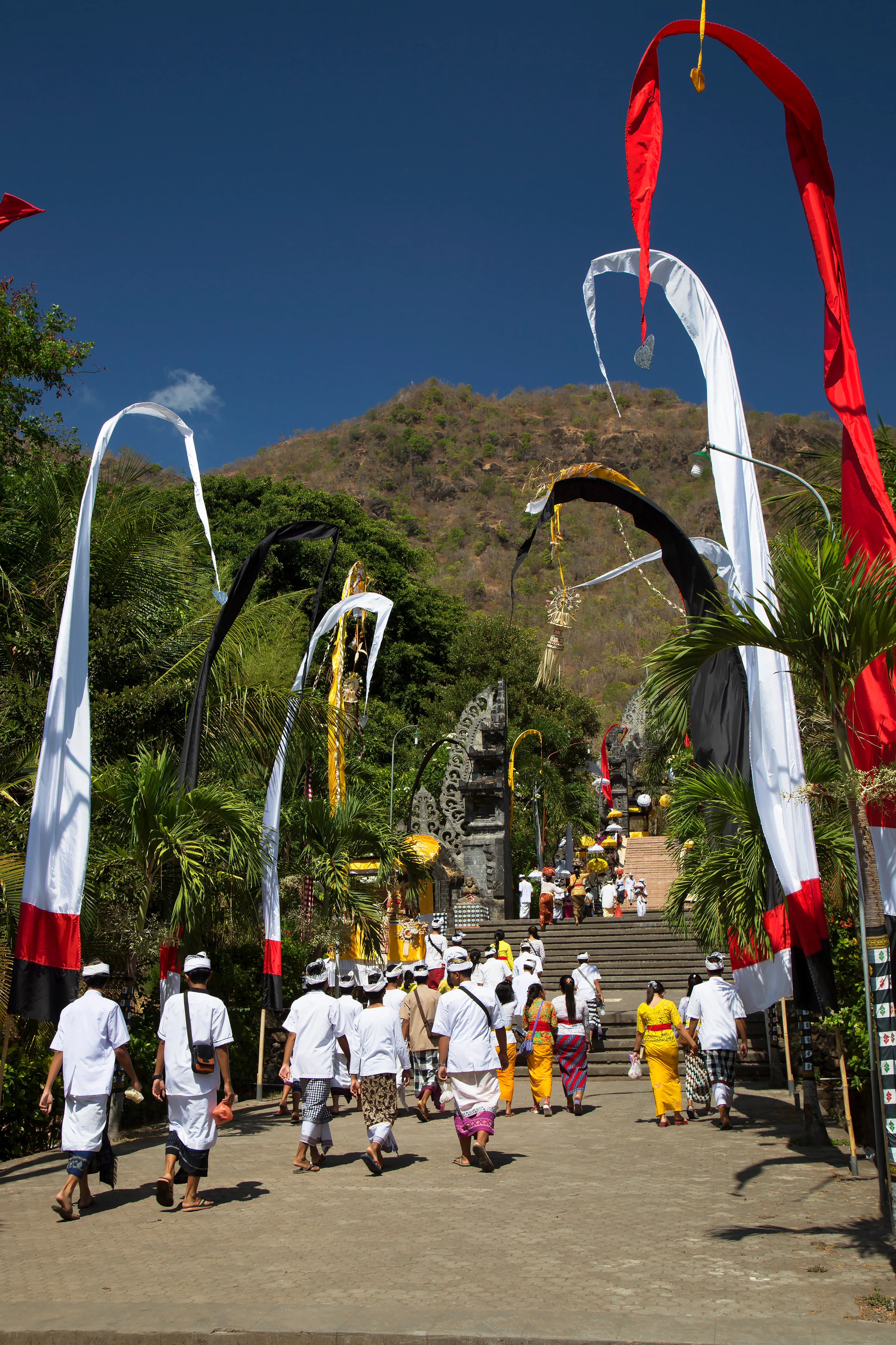 People in white walking up temple steps lined with tall penjor poles in Bali