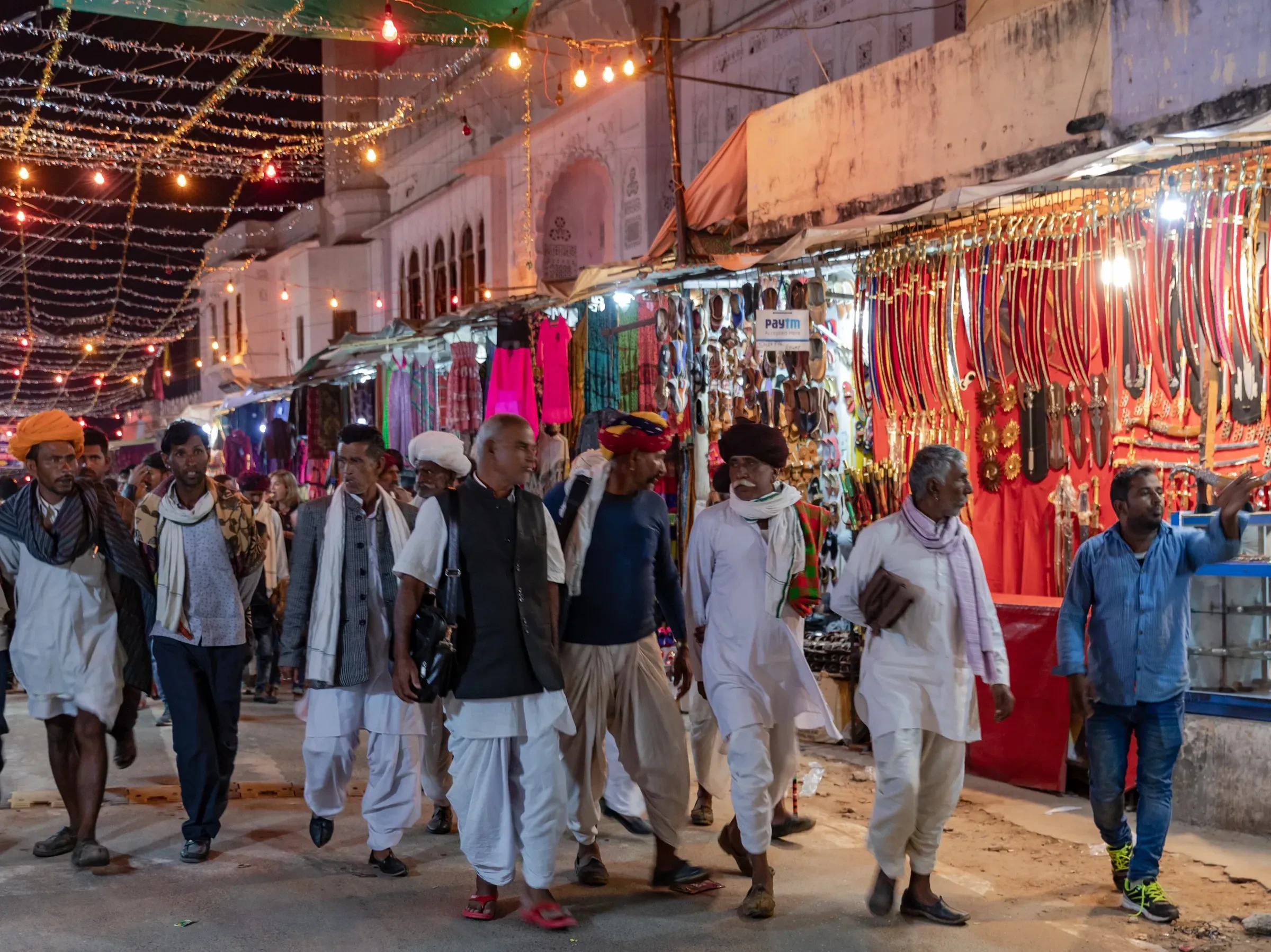 Men in turbans walking past lit stalls in the Pushkar fair bazaar in the evening