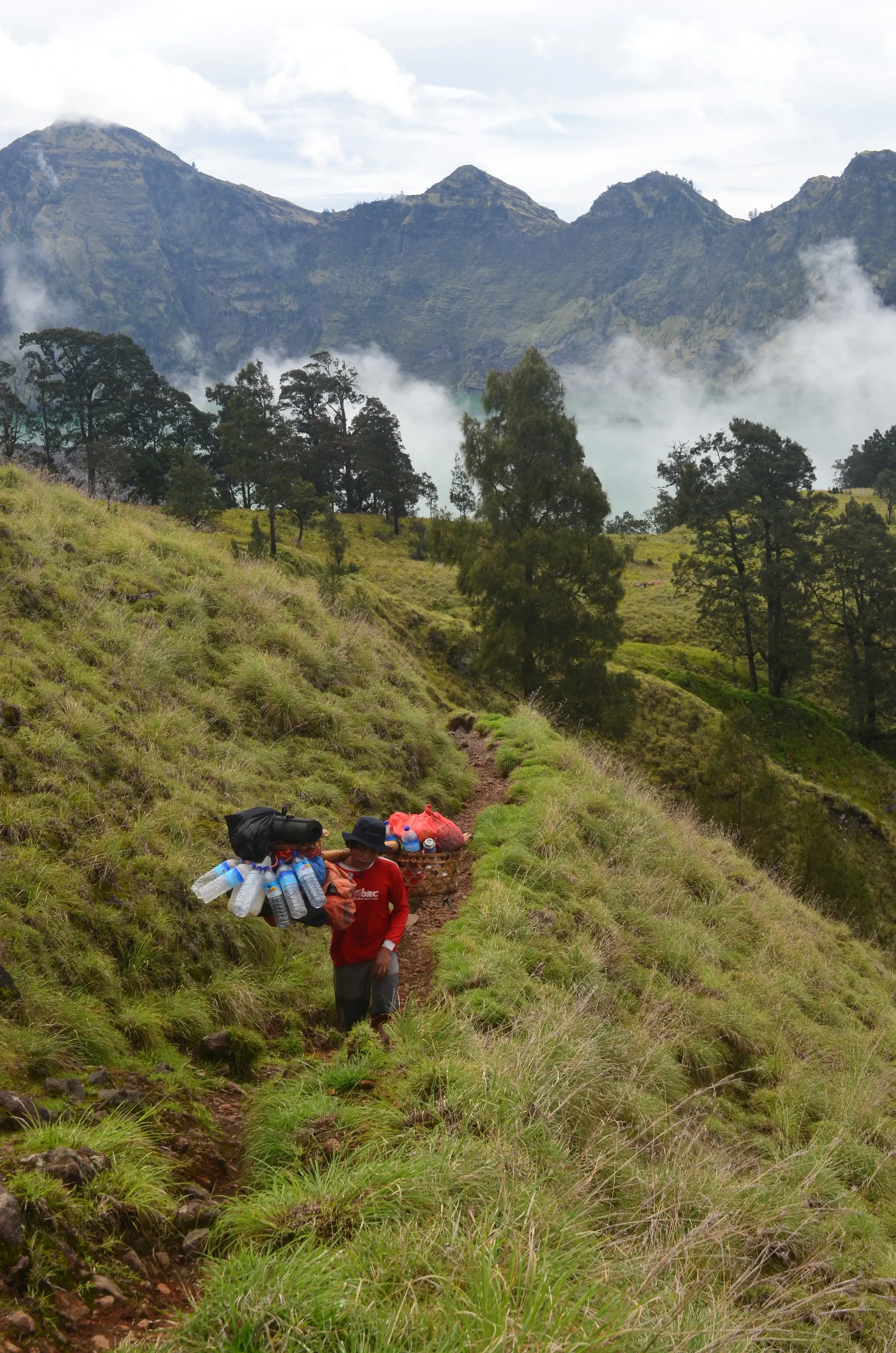 A porter carrying a heavy load up a grassy Rinjani trail, with ridges and cloud behind