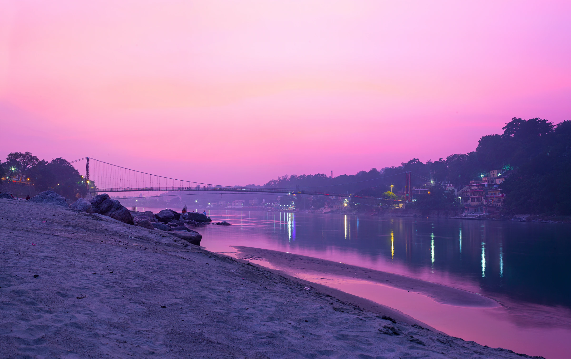 A suspension footbridge over the Ganga at dusk, with lights along the riverbank in Rishikesh