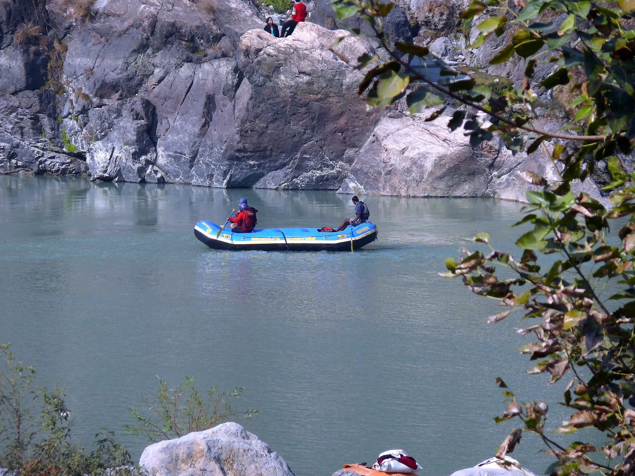 A rafting crew in helmets and life jackets on a calm teal stretch of the Ganga near Rishikesh