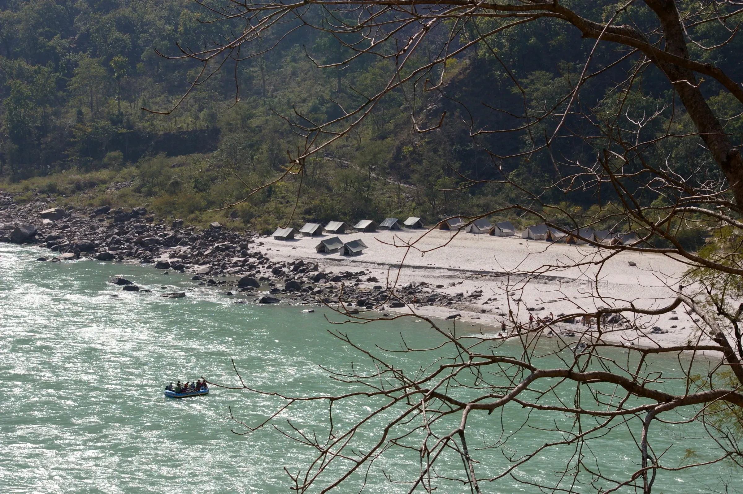 A row of tents in a riverside camp on a sandy Ganga beach, with a raft on the water and forested hills behind