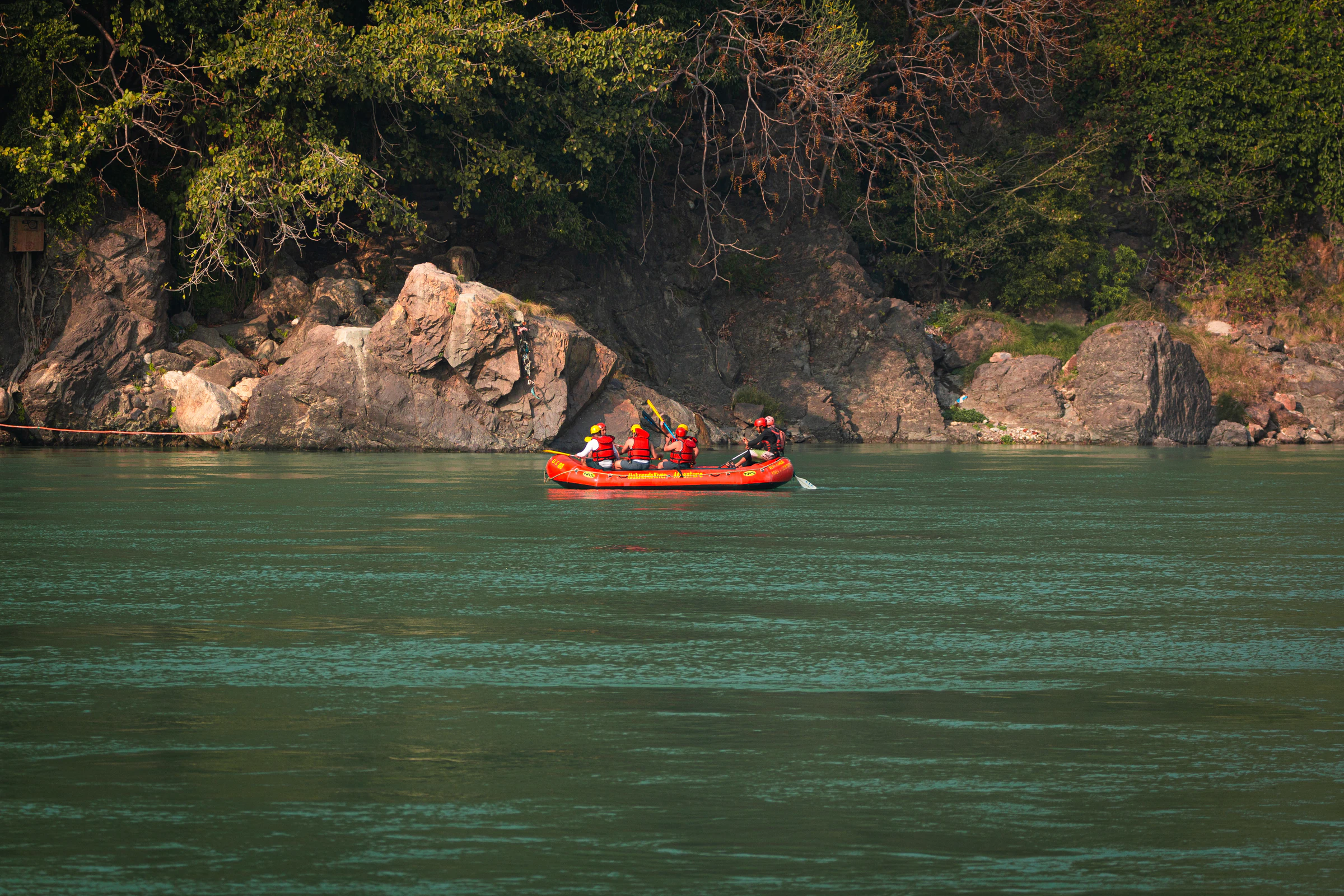 A raft crew in helmets and life jackets on the green Ganga near Rishikesh, with a rock wall behind