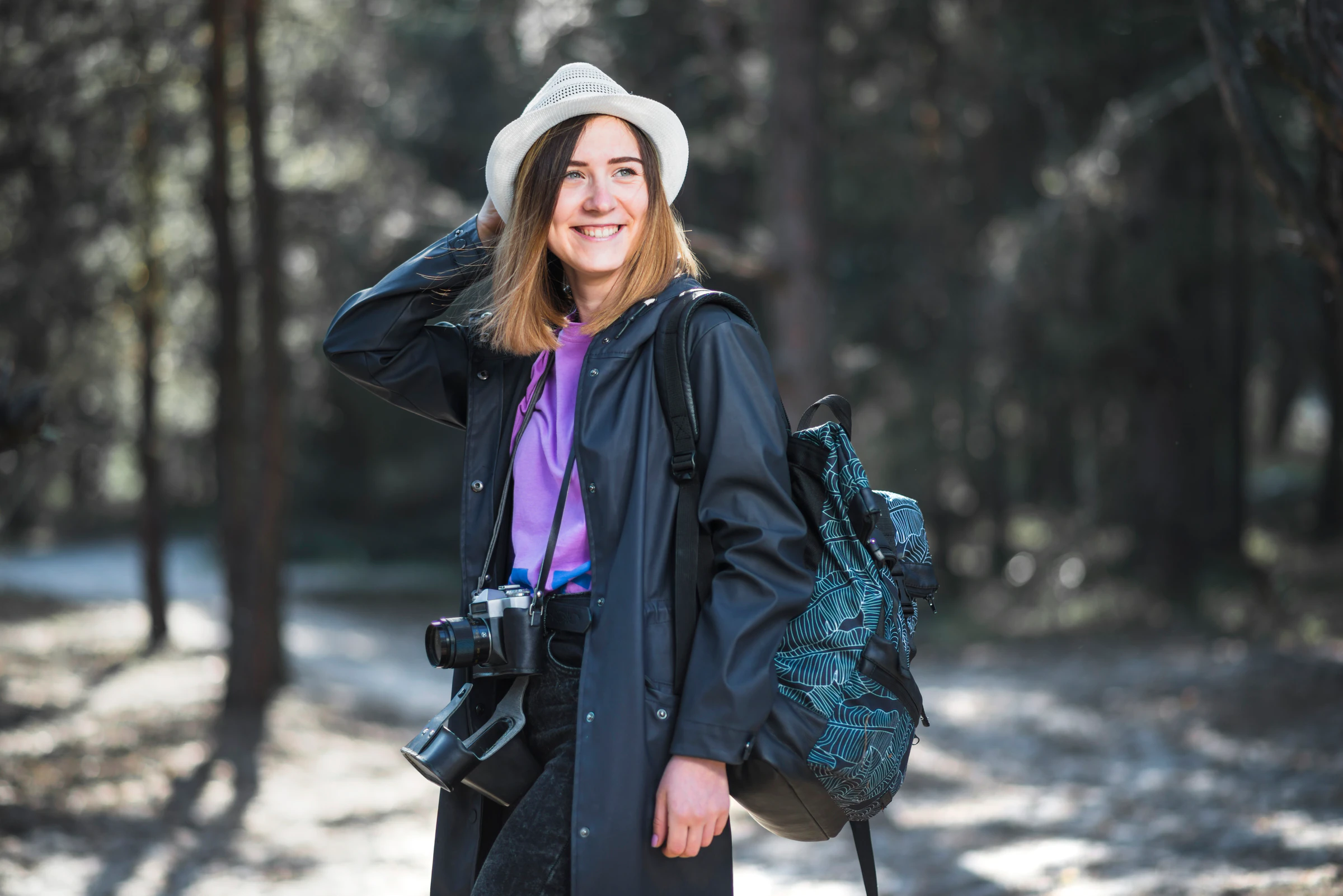A smiling woman with a backpack and camera standing on a forest path