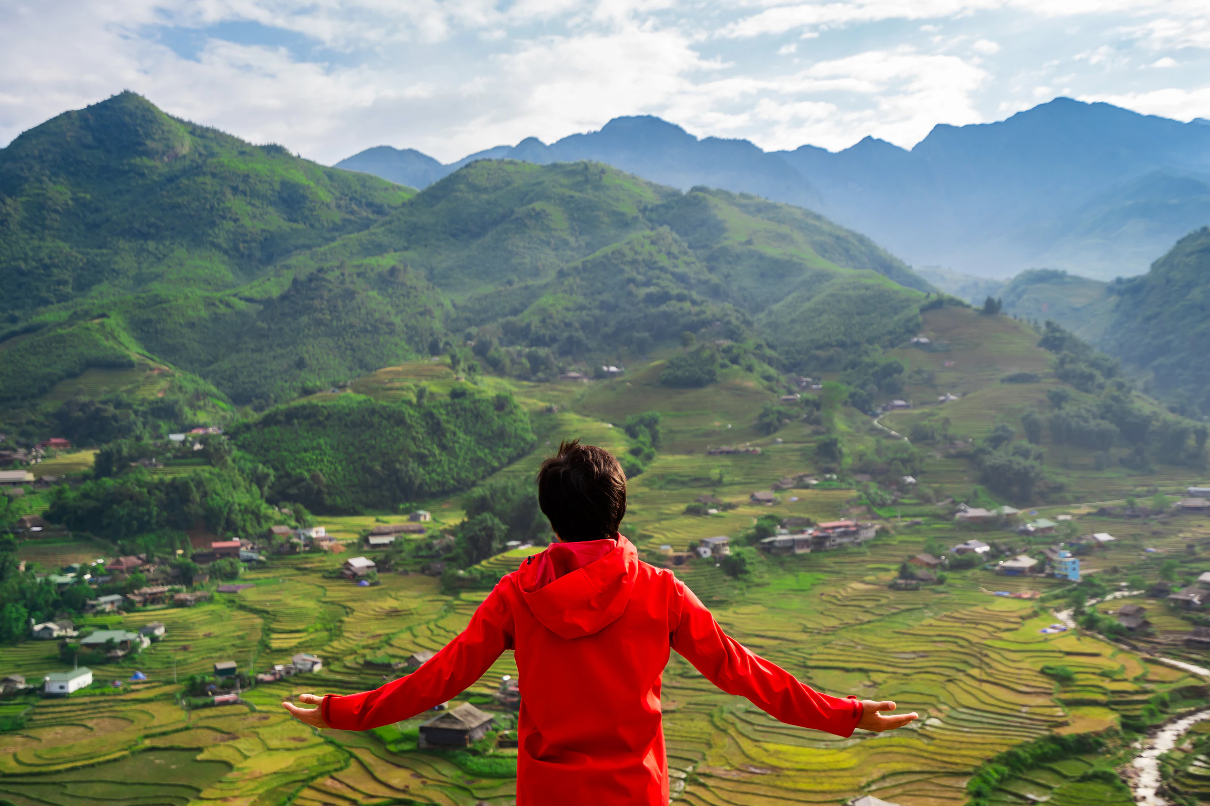 A traveller in a red jacket stands with arms outstretched above green rice terraces and villages in Sapa