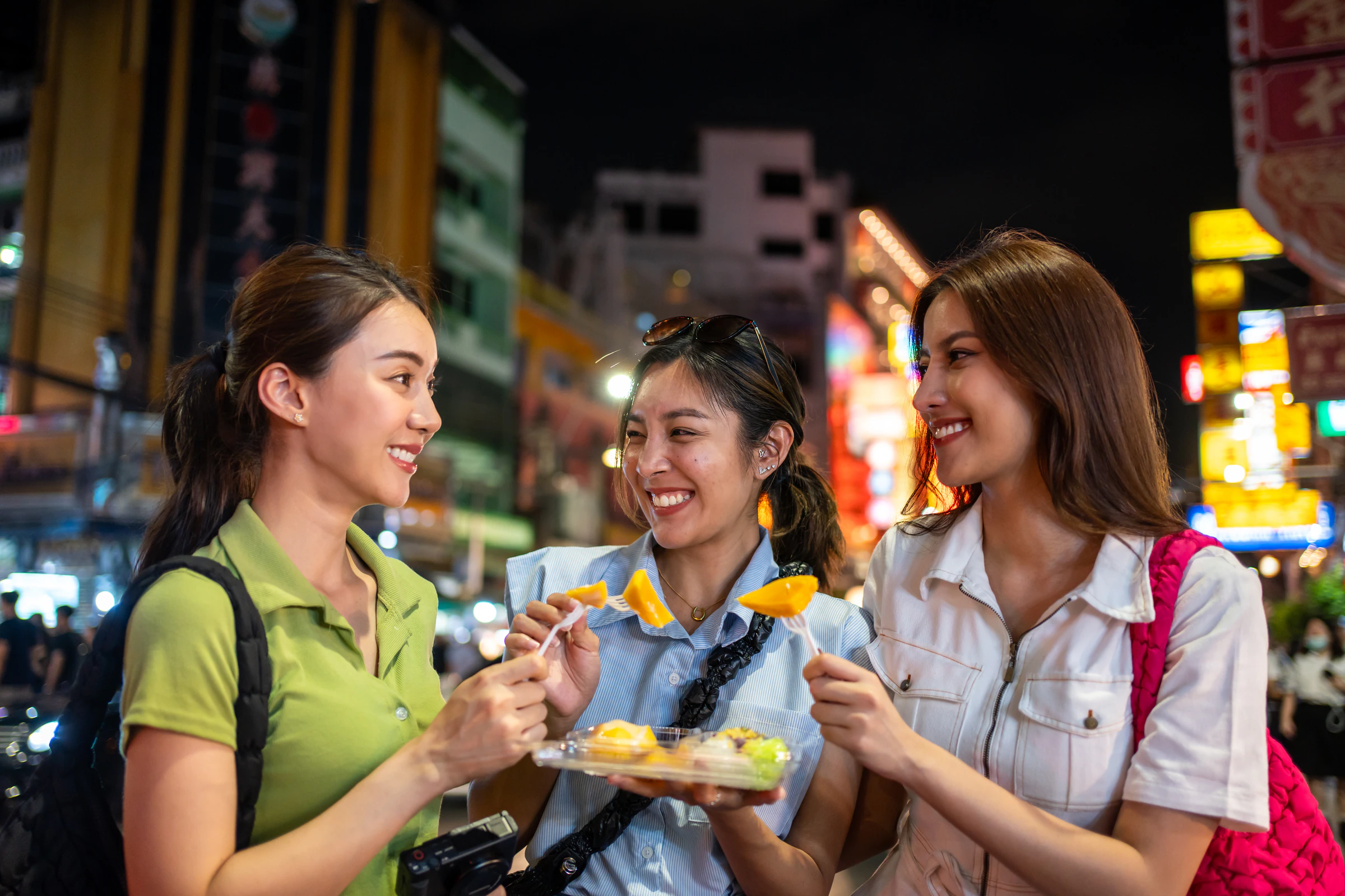 Three women friends laughing and sharing street food on a busy street at night