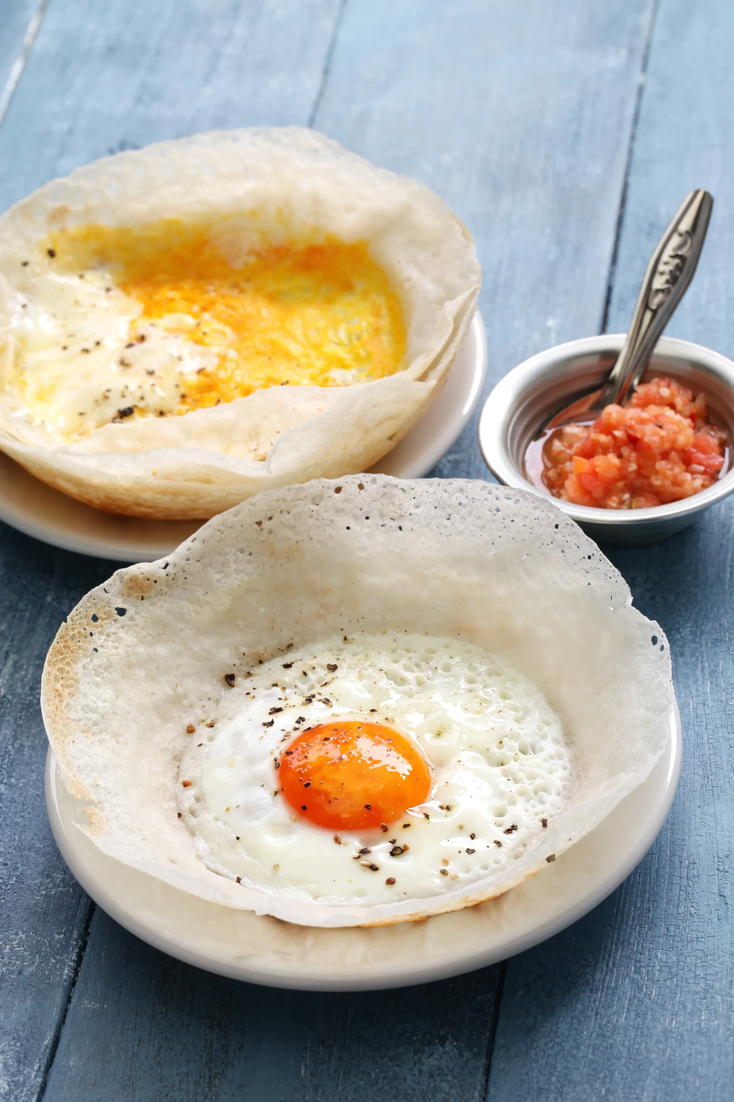 Two egg hoppers with a small bowl of red salsa on a blue wooden table