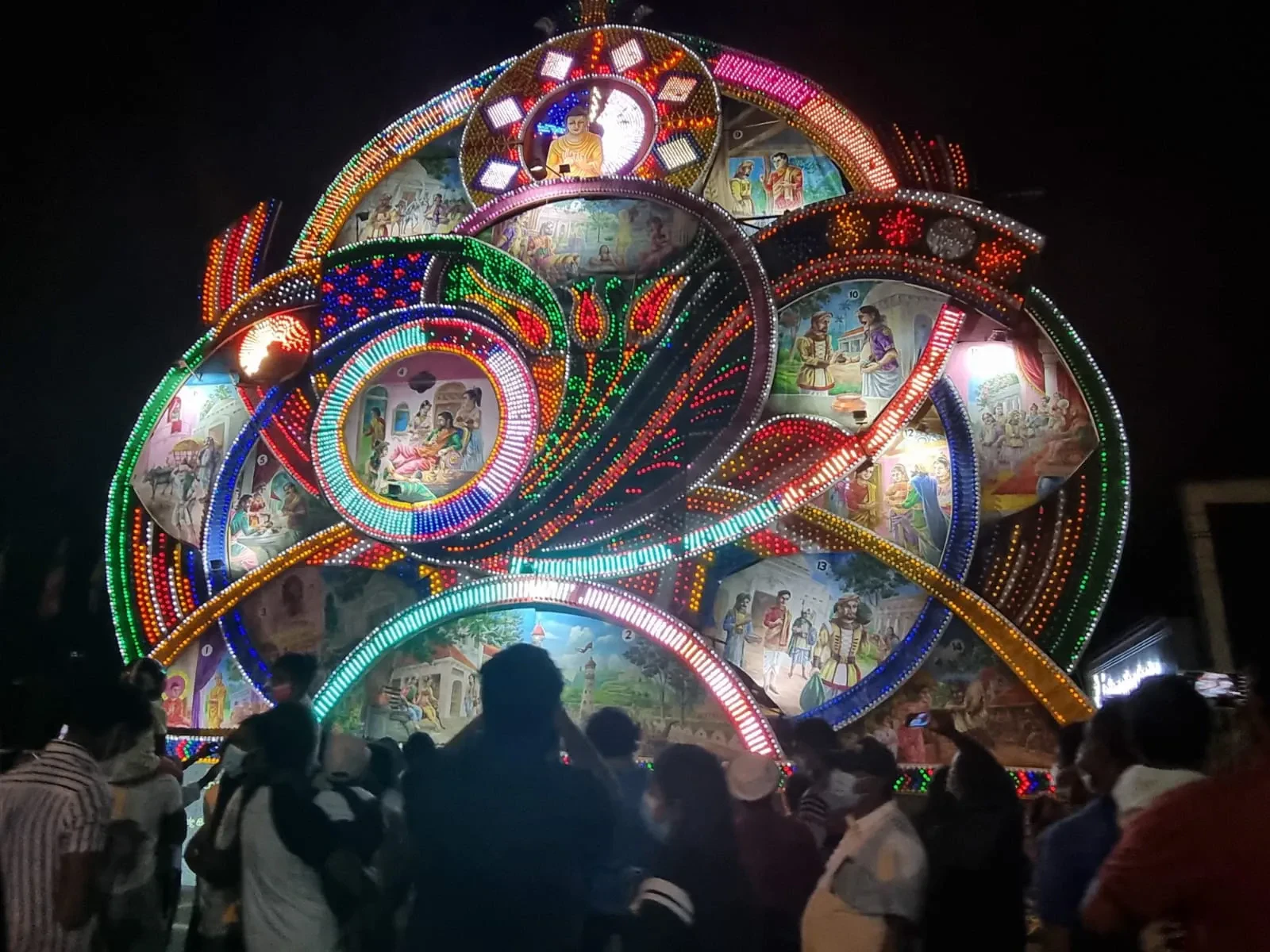 A brightly lit Vesak pandol with painted Buddhist story panels at night in Maharagama