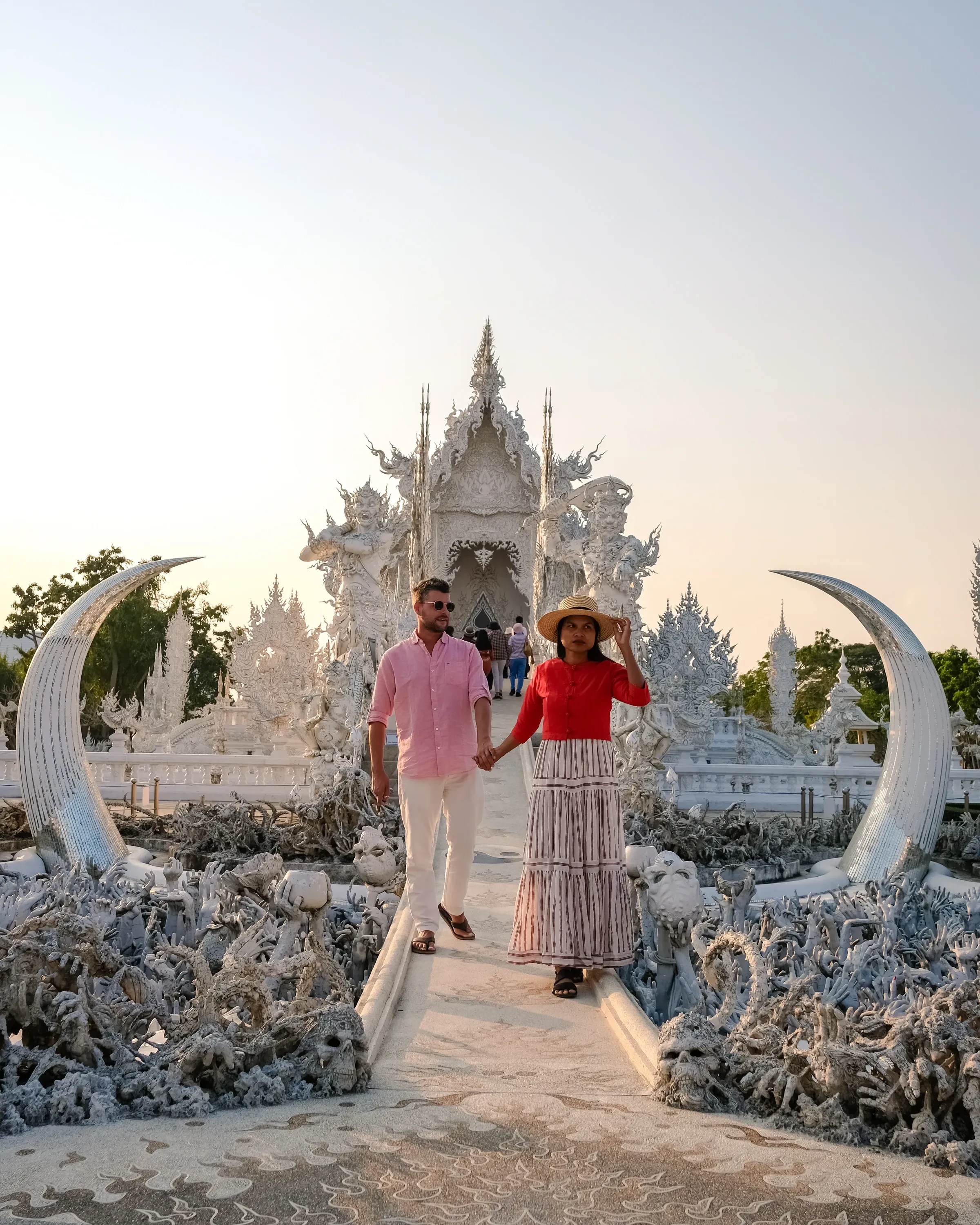 A couple walking across the bridge towards the carved white temple of Wat Rong Khun in Chiang Rai in warm evening light