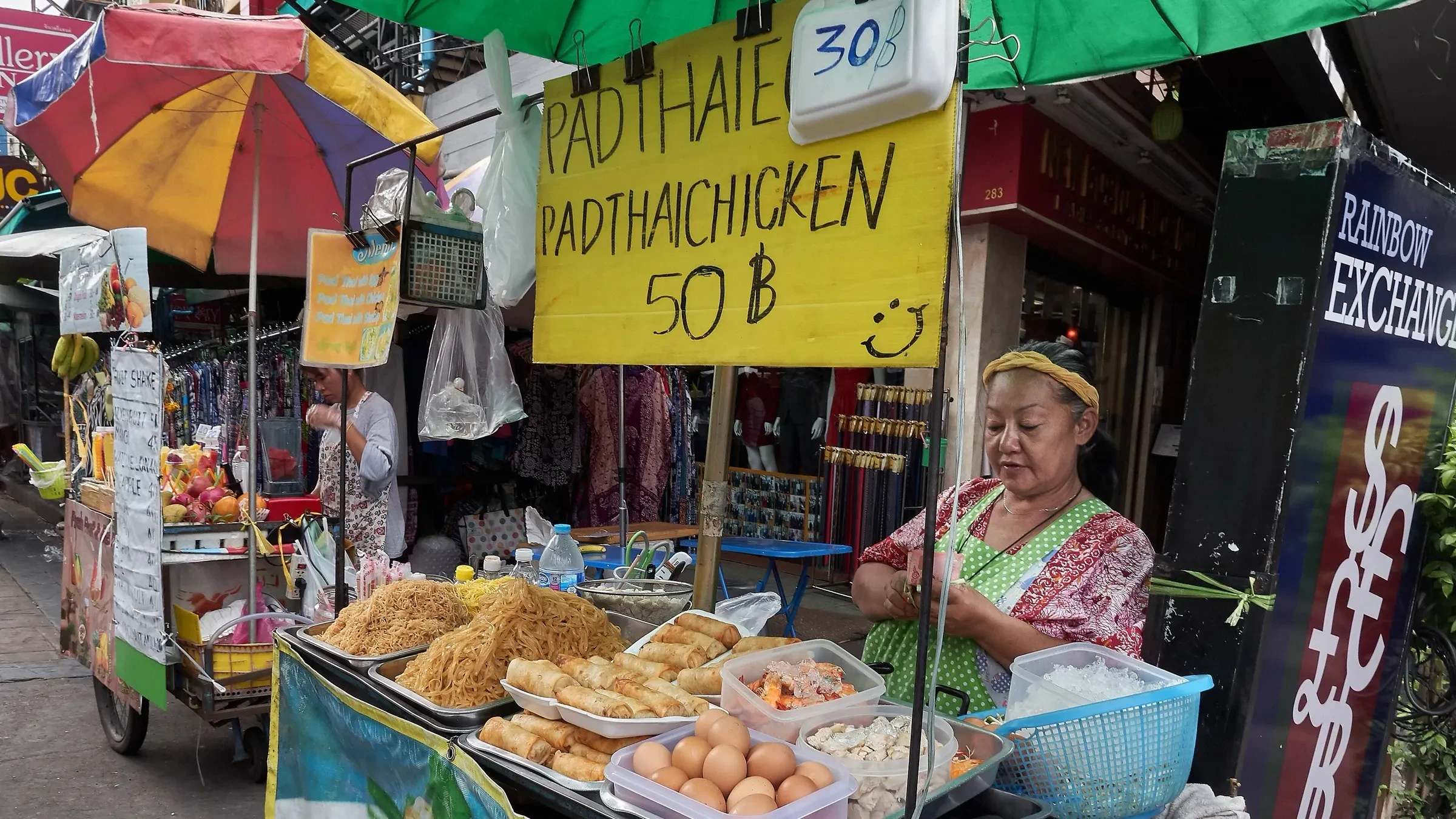 A street vendor selling pad thai and fried snacks at a stall with a price sign in Bangkok