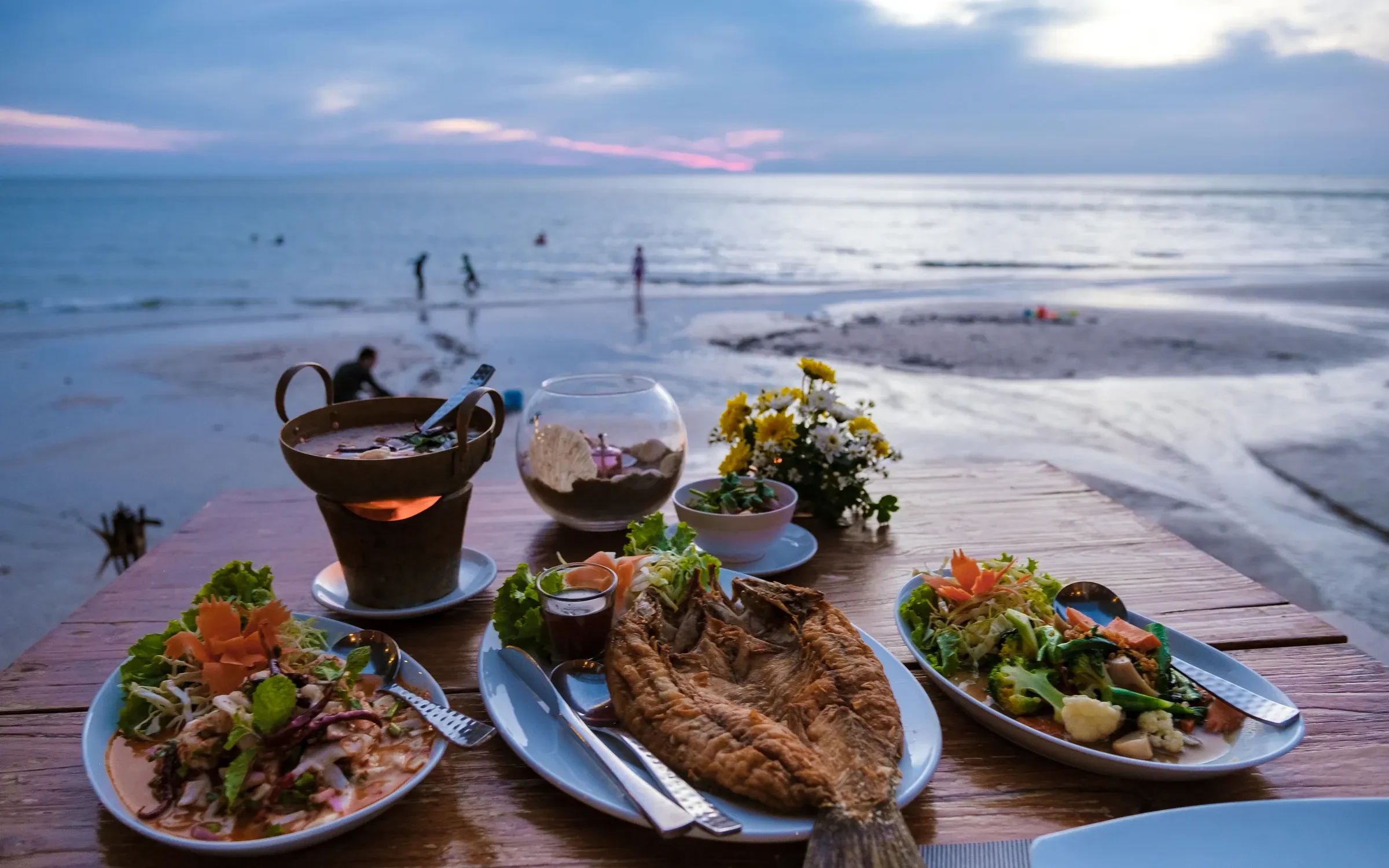 Grilled fish and Thai dishes laid out on a beachfront table at sunset
