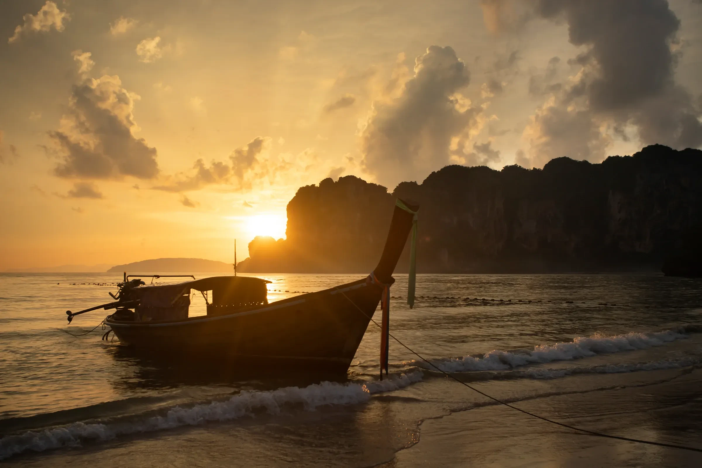 Sunset over Railay beach in Krabi, with a longtail boat silhouetted against the cliffs