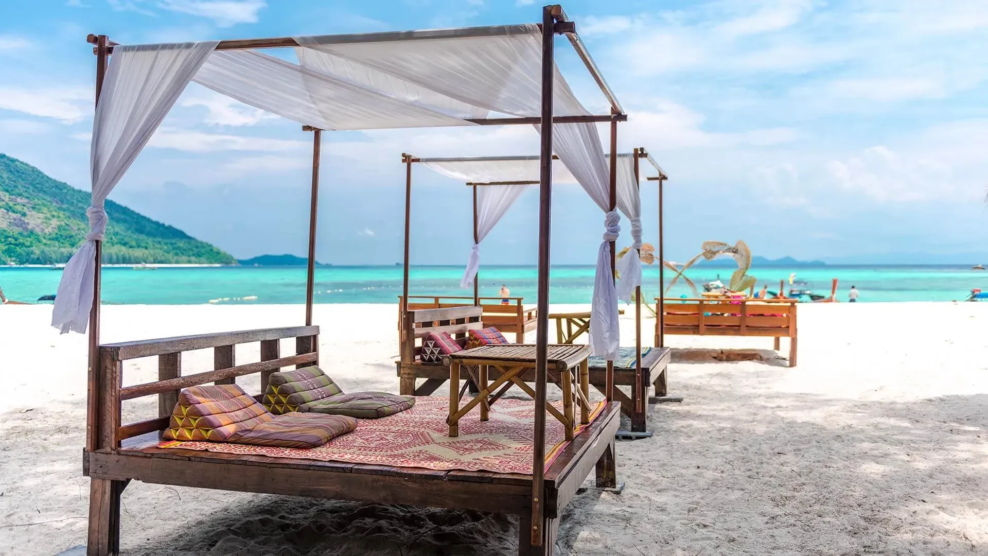 White-draped wooden daybeds on Sunrise Beach with turquoise water behind, Koh Lipe