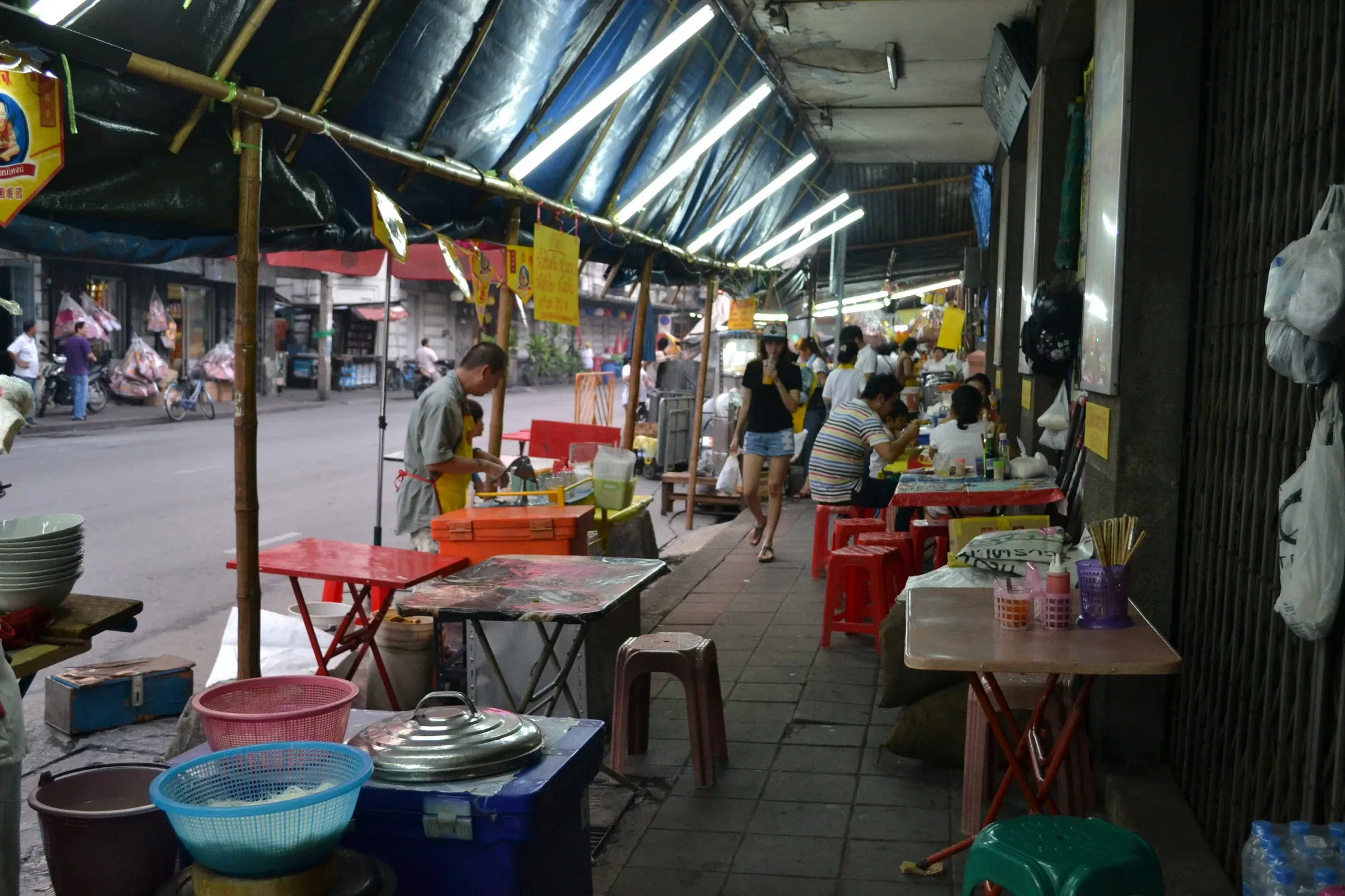 A Bangkok street lined with food stalls under tarpaulins, with people eating at small tables