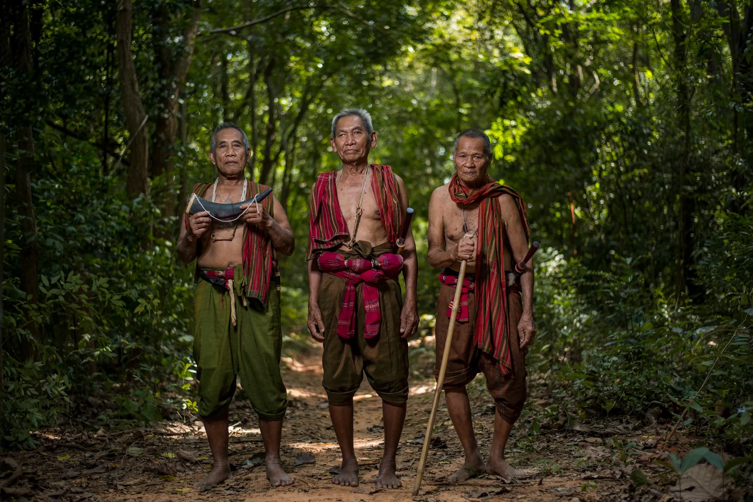 Three older mahouts in traditional dress standing on a forest path in Surin province