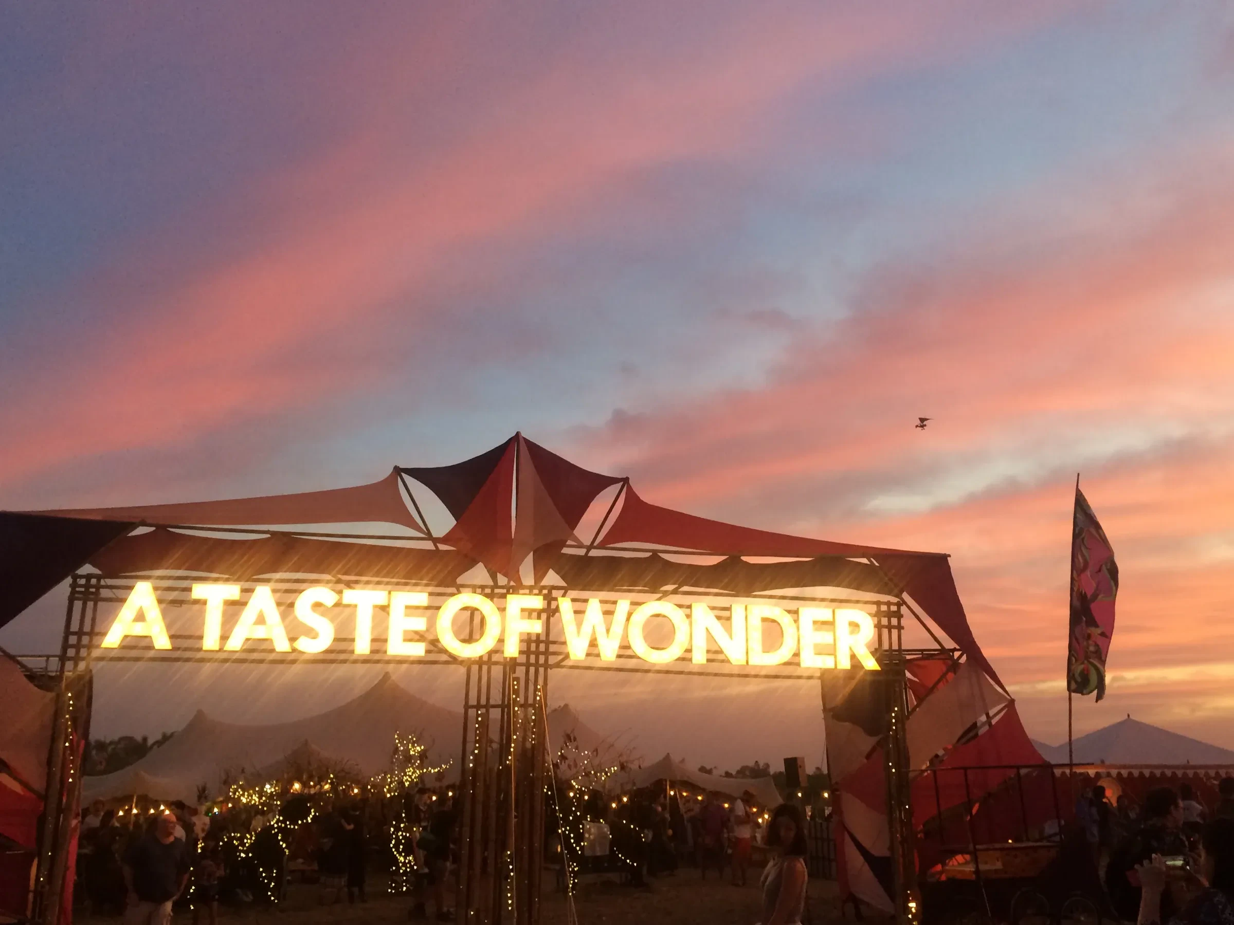 The lit \"A Taste of Wonder\" gate against a pink sunset sky at Wonderfruit