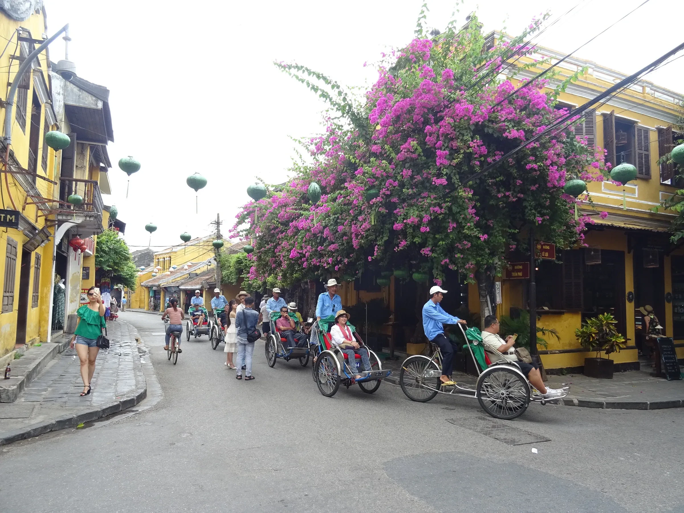 Cyclos and cyclists on a street of yellow shophouses under a flowering bougainvillea in Hoi An